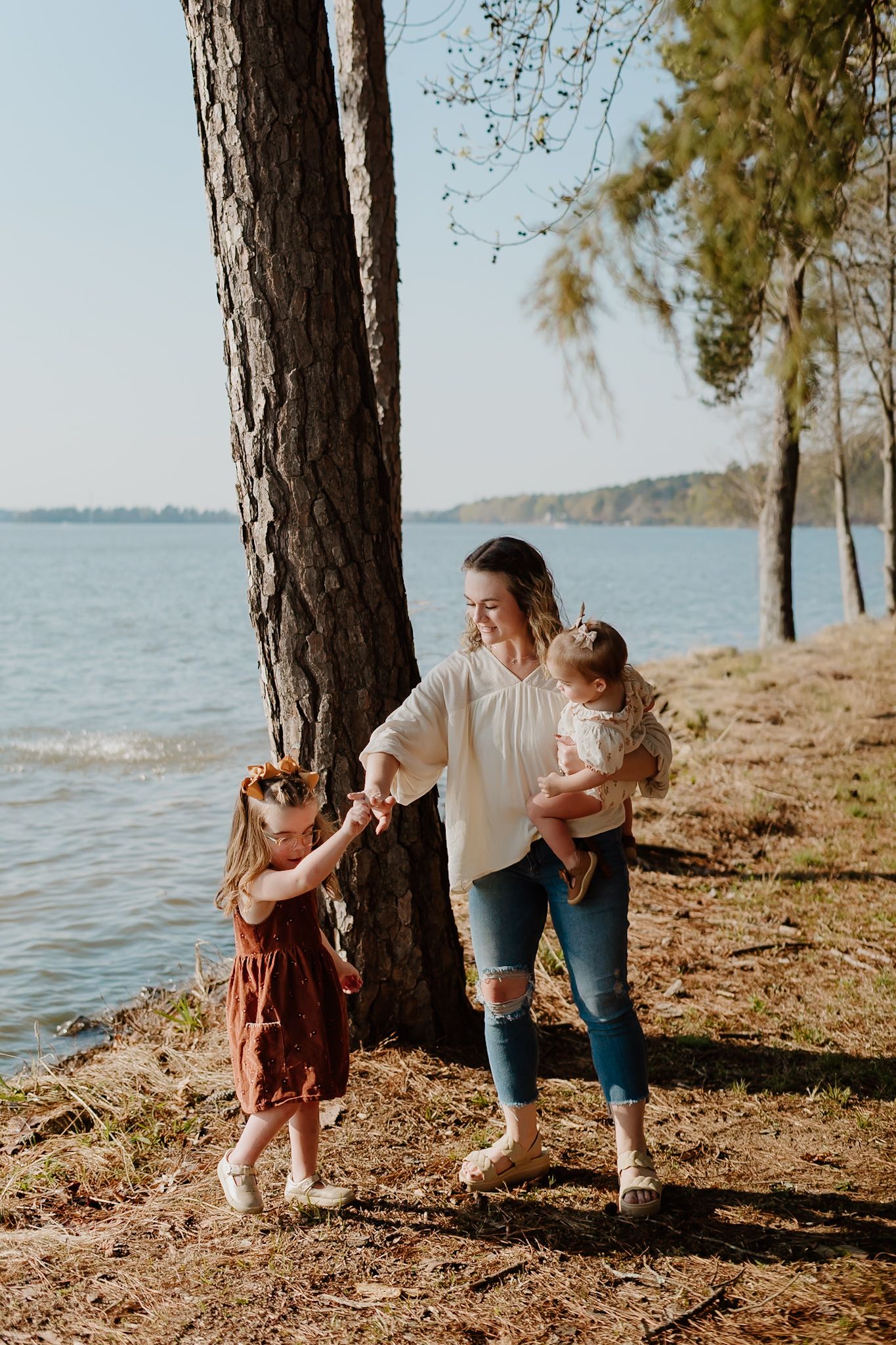 A mother holds her baby while dancing with her young daughter near a lakeside pine tree on a sunny day.
