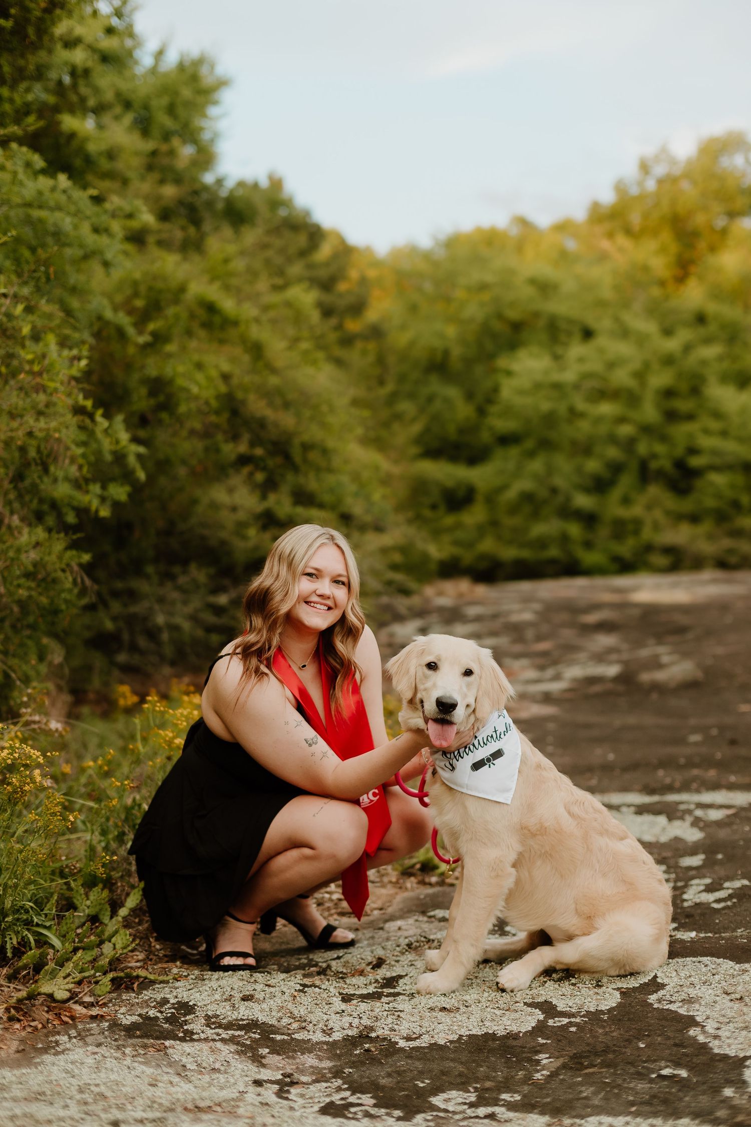 Smiling blonde woman in red top crouches beside a golden retriever wearing a white bandana on a rocky outdoor path.