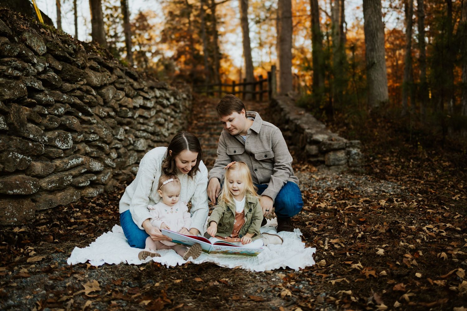 Family of four reading book together on white blanket surrounded by fall foliage outdoors.