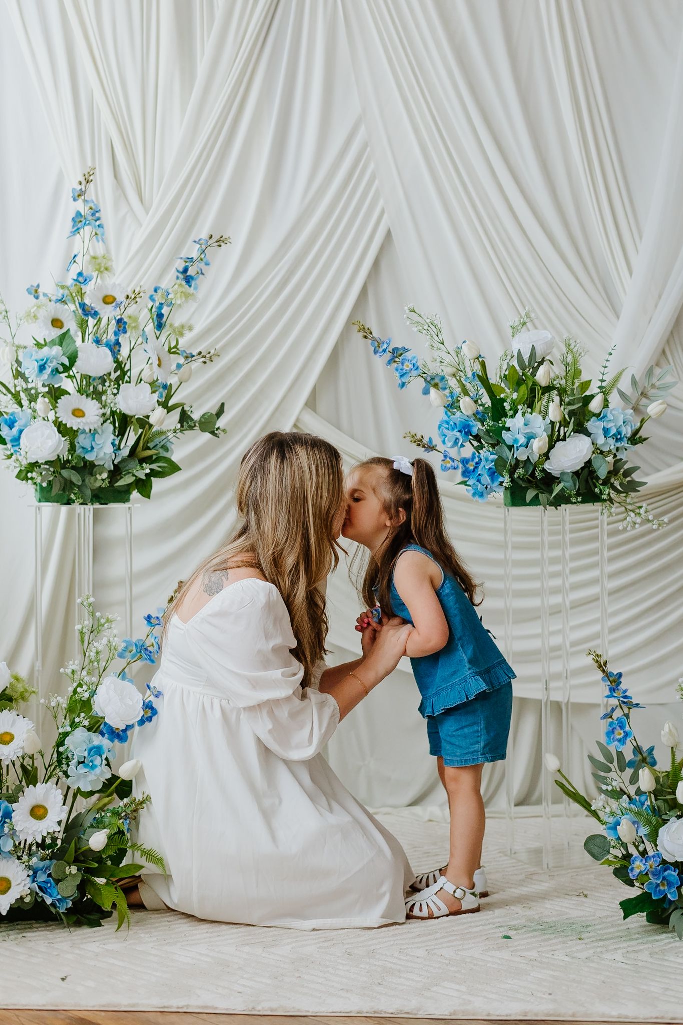 A mother in a white dress shares a kiss with her young daughter in a blue outfit amid elegant floral decor.