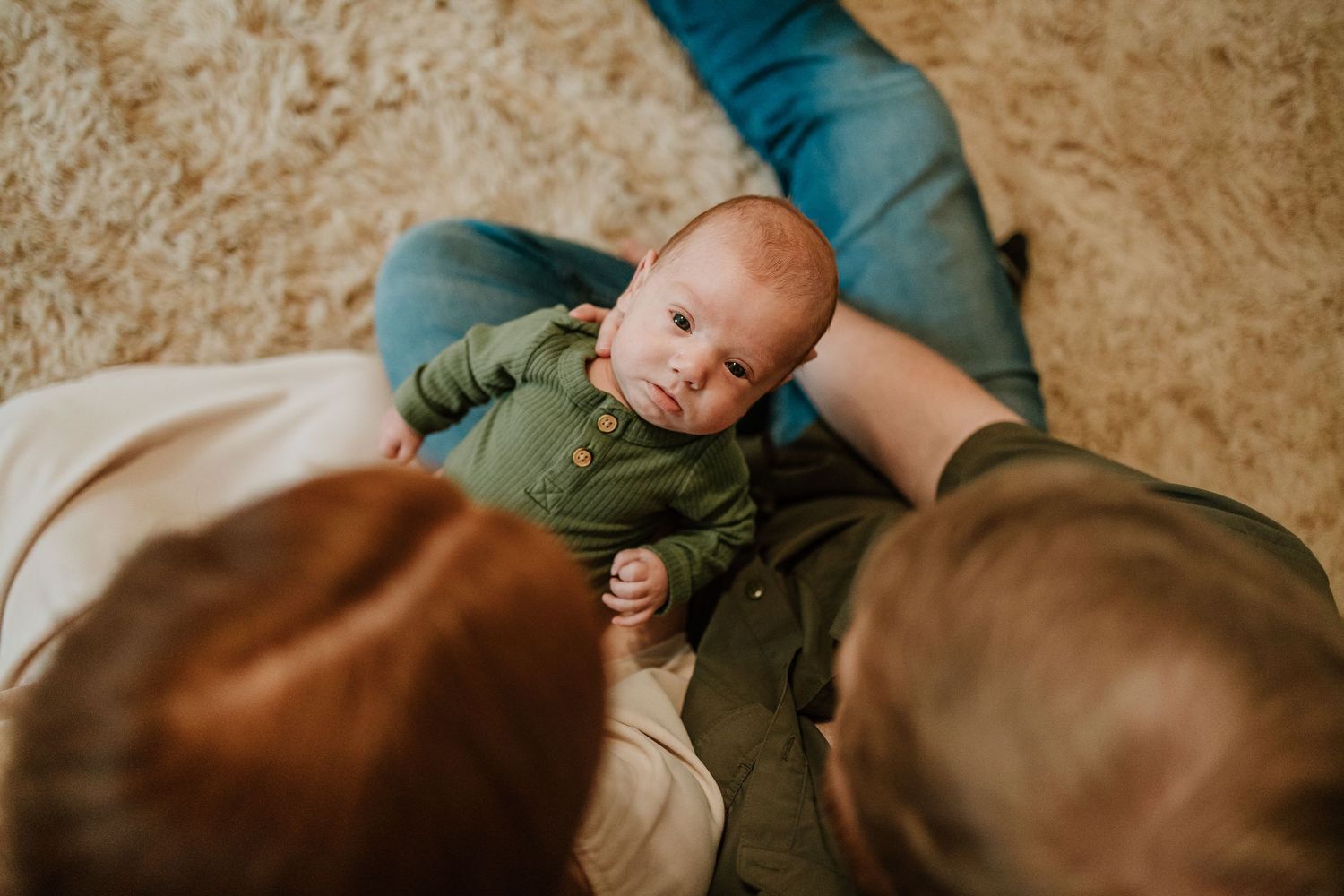Overhead view of new parents cradling their newborn baby in a green onesie on a fluffy rug.