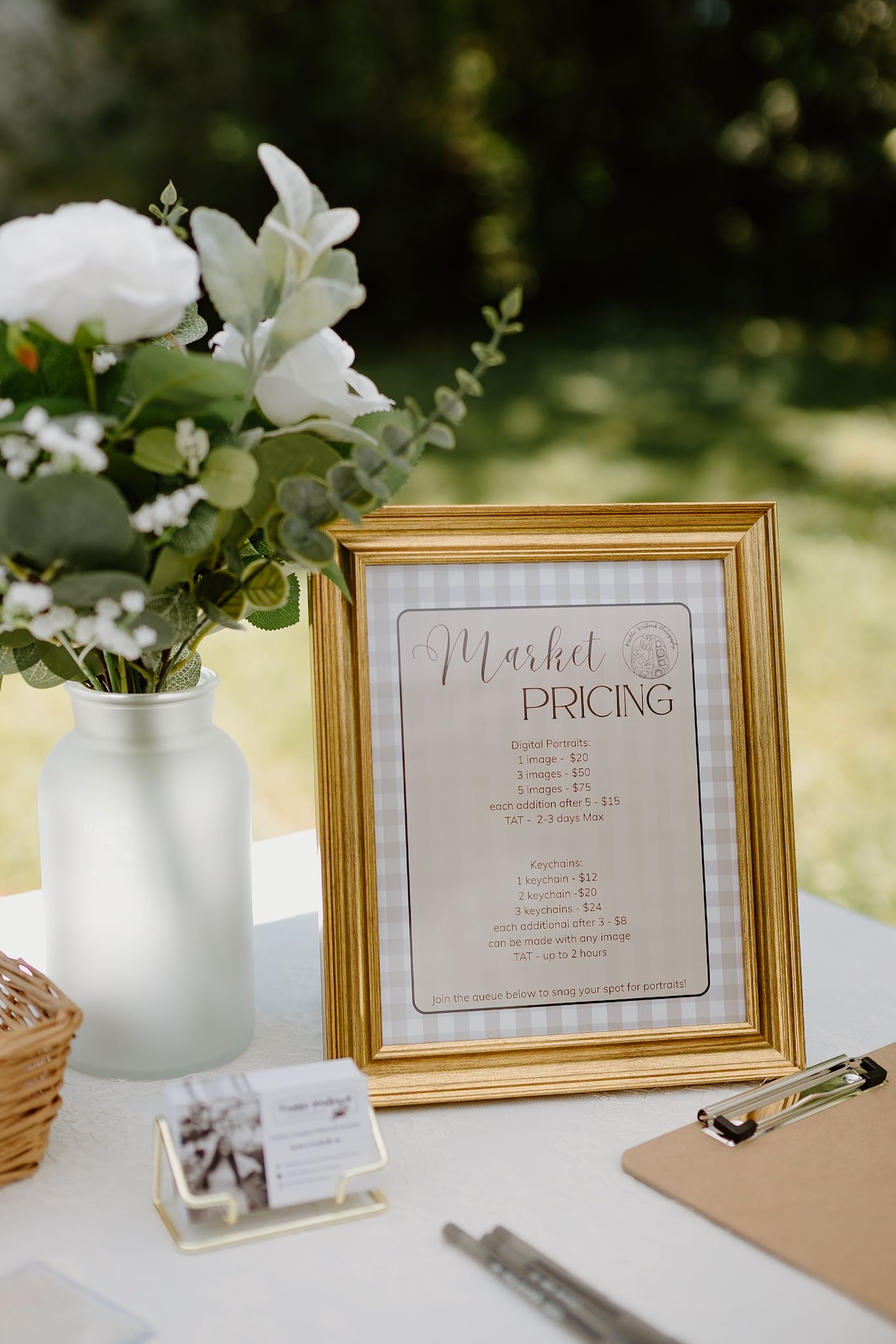 Gold framed market pricing sign on white table with white floral arrangement, clipboard, and business cards at outdoor event.