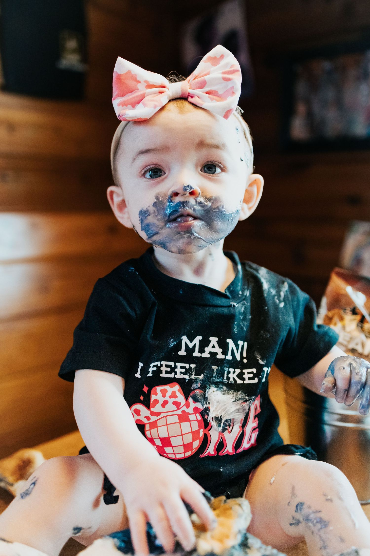 Messy baby girl with cake-covered face in first birthday shirt during fun cake smash photoshoot.