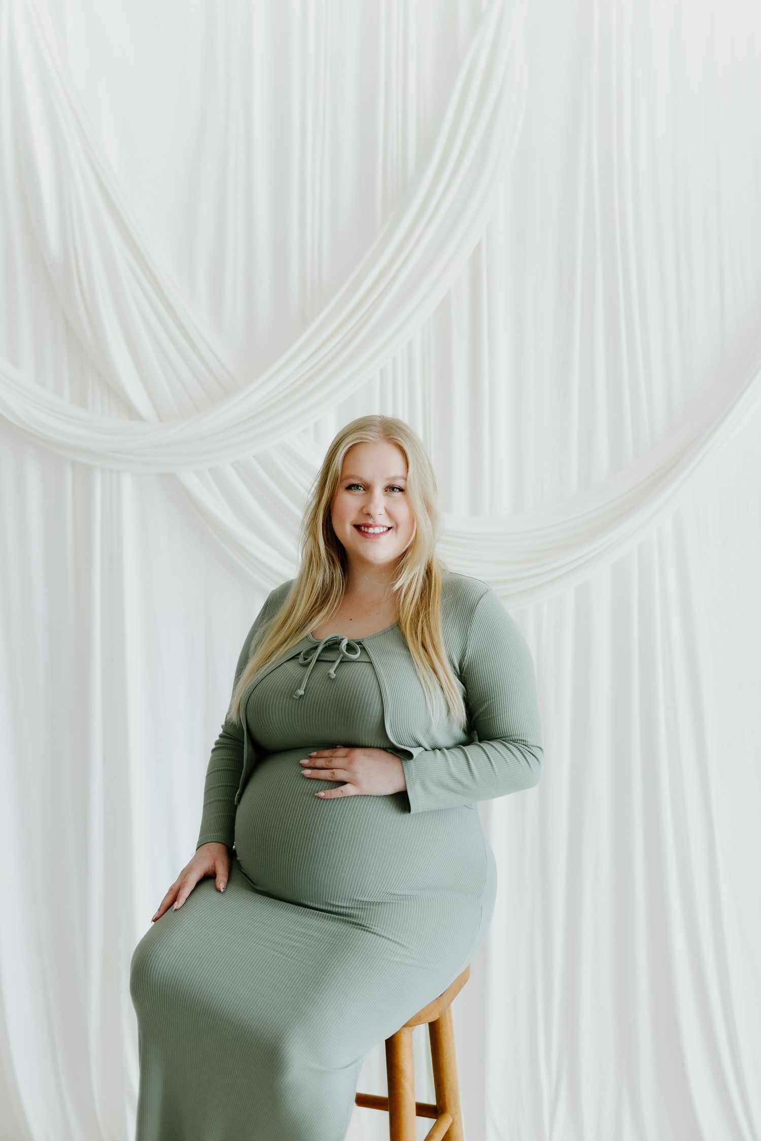 Radiant pregnant woman in sage ribbed maternity dress sits on stool against white draped backdrop.