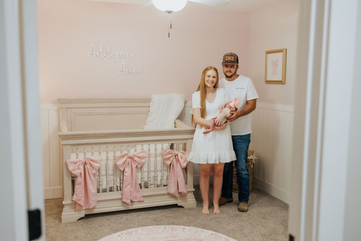 New parents stand proudly in a pink nursery holding their newborn beside a white crib adorned with pink bows.