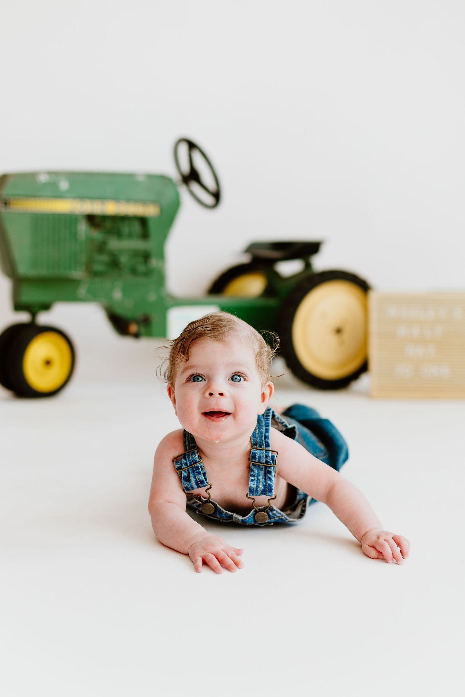 Happy baby in denim overalls doing tummy time near John Deere tractor prop.