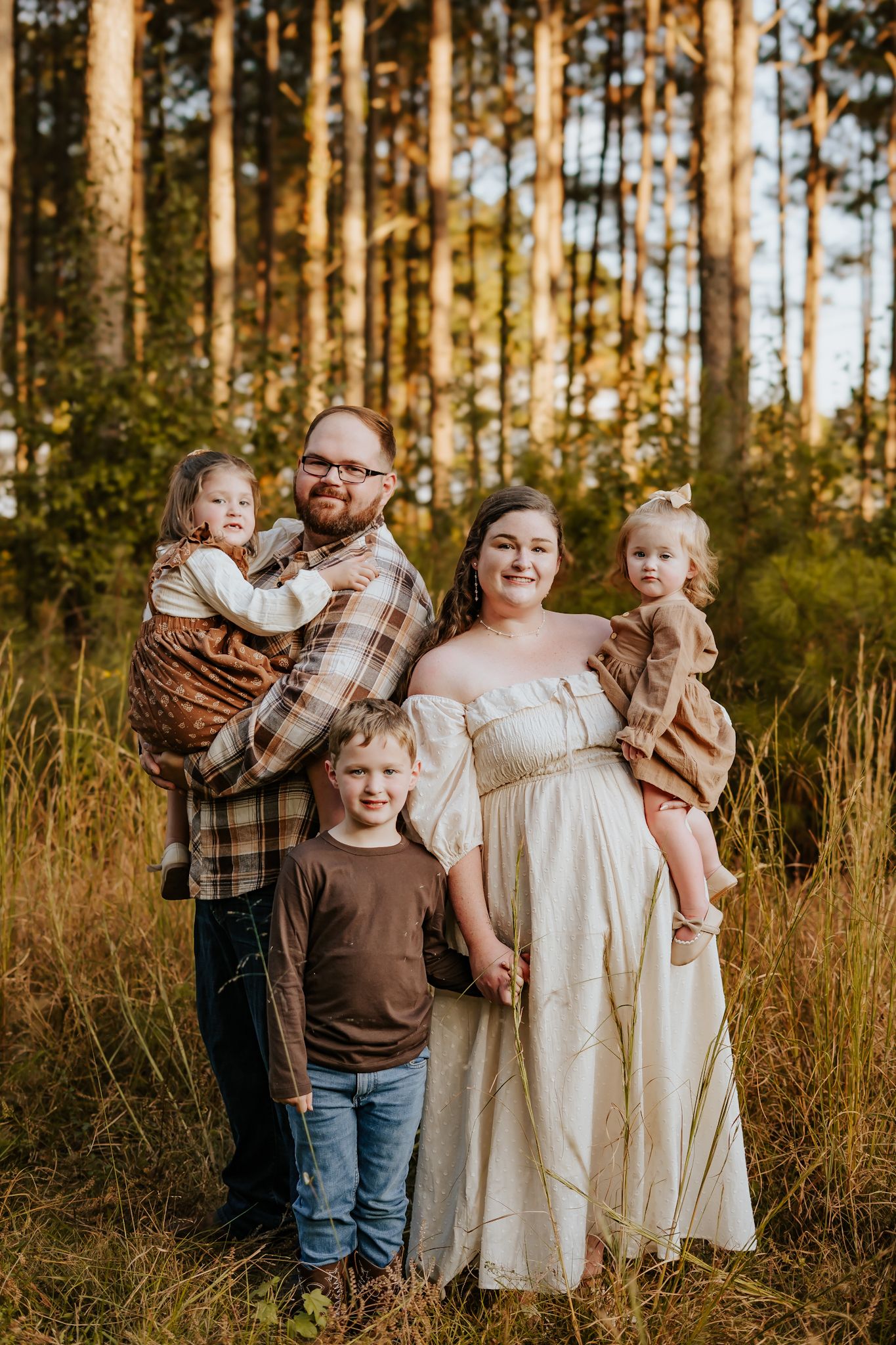 Family of five posing together in a golden pine forest at sunset, mother in a flowing white dress.