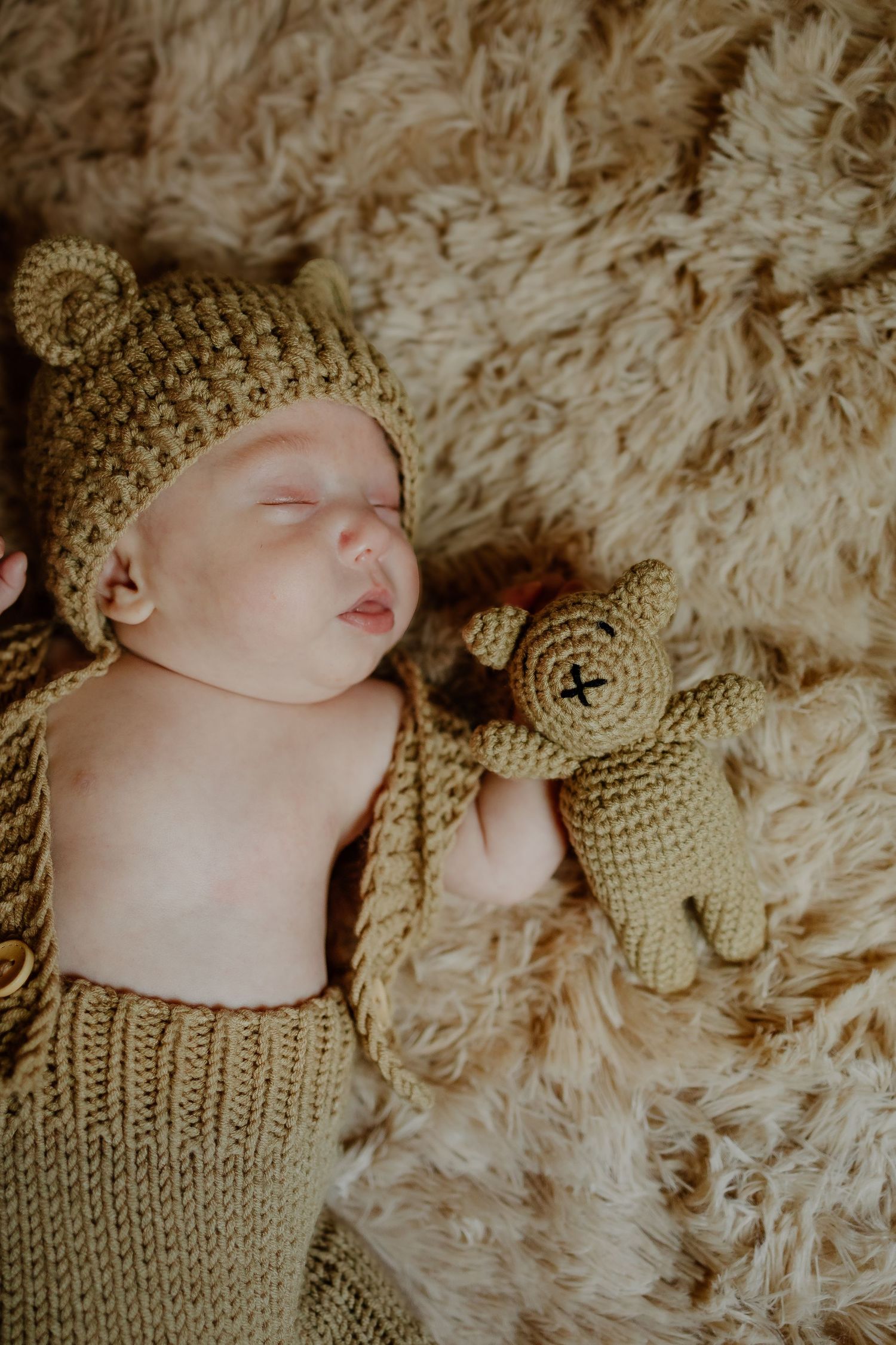 Sleeping newborn baby wearing a knit bear hat beside a matching crochet teddy bear on fur.