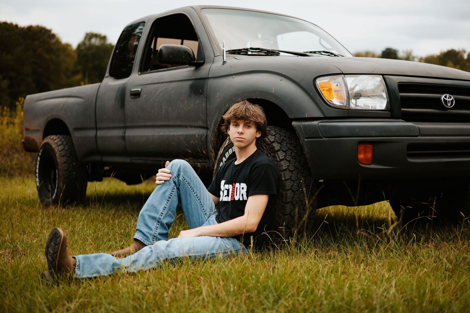 Teen boy in black senior shirt sitting in grass leaning against matte black Toyota Tacoma truck.