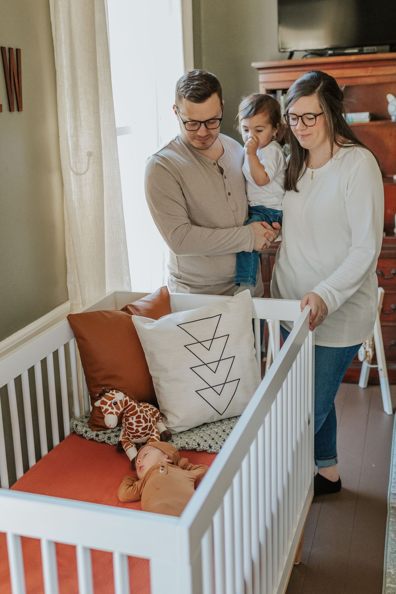 Young family with toddler and parents stand together admiring a white baby crib in nursery.