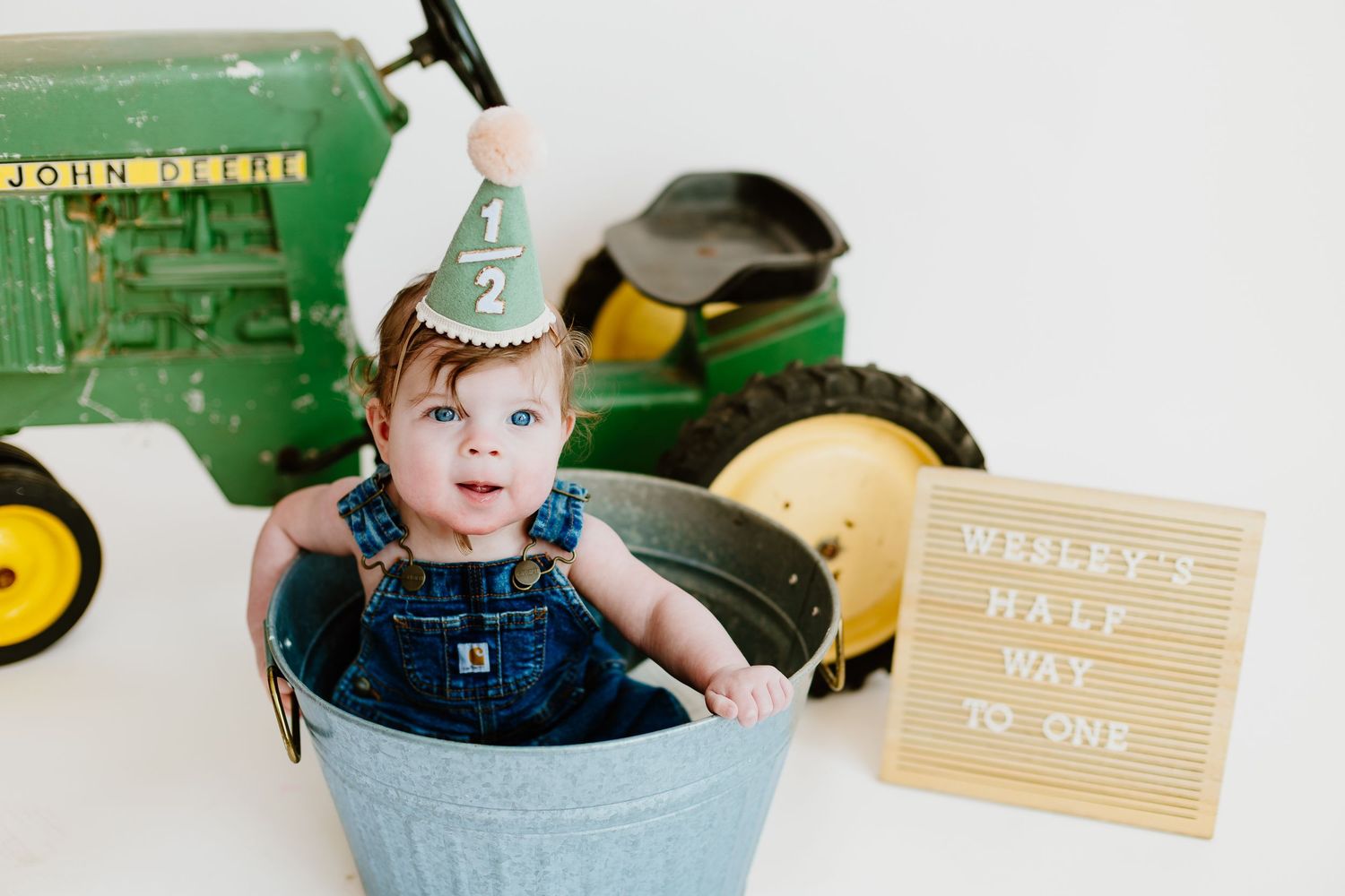 Baby in overalls wearing half birthday hat in bucket beside John Deere tractor and letter board.