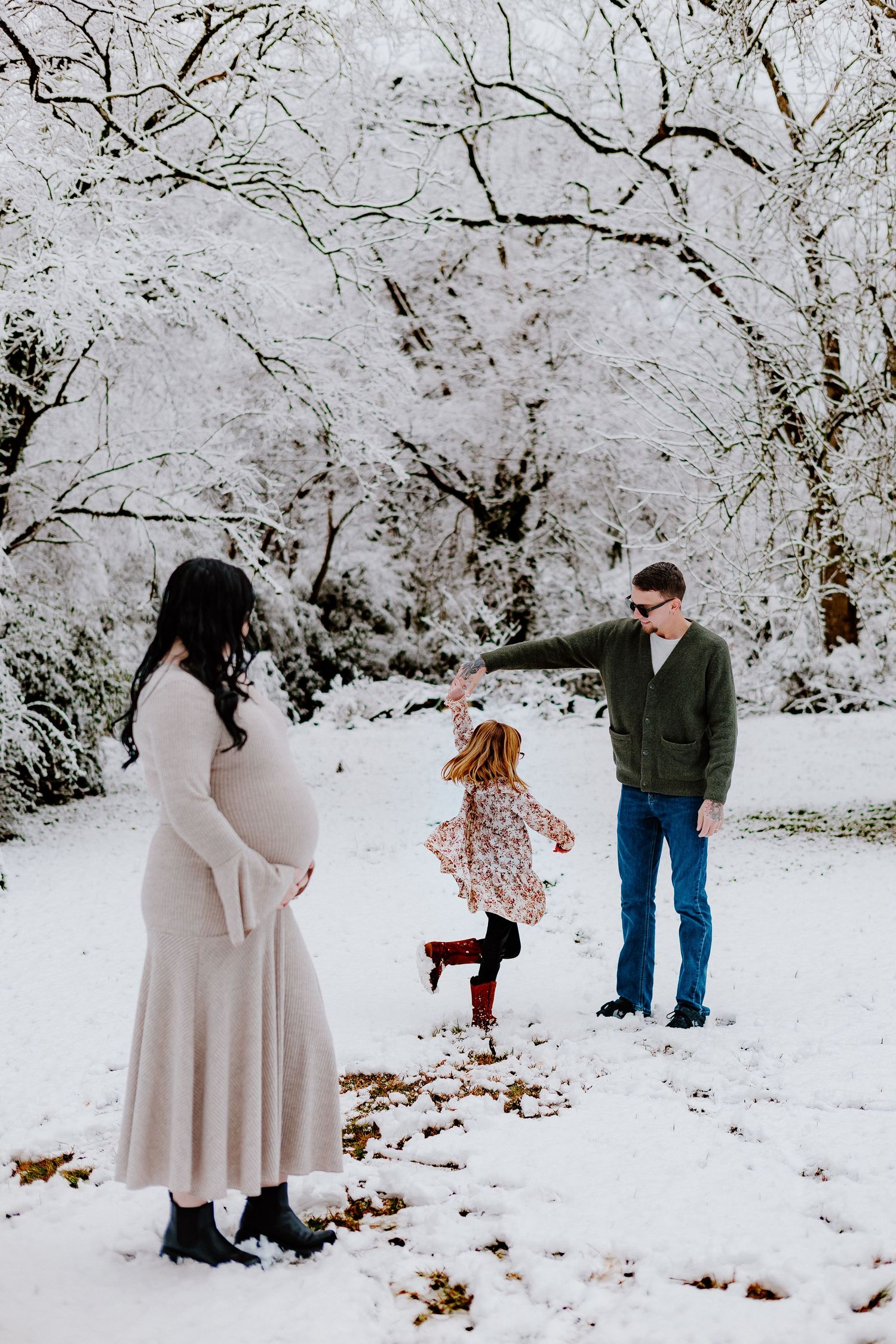 Family maternity photo with couple and toddler playing in snow-covered winter park.