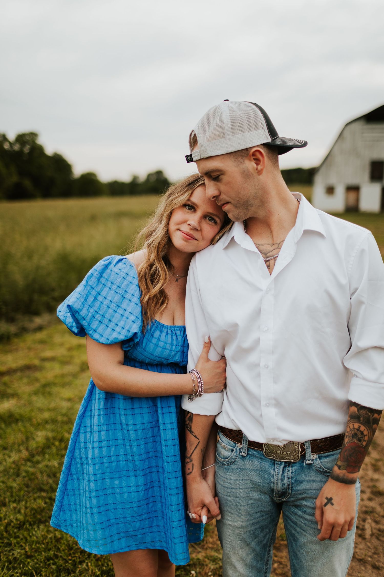 Woman in blue dress leaning on tattooed man in white shirt near a barn.