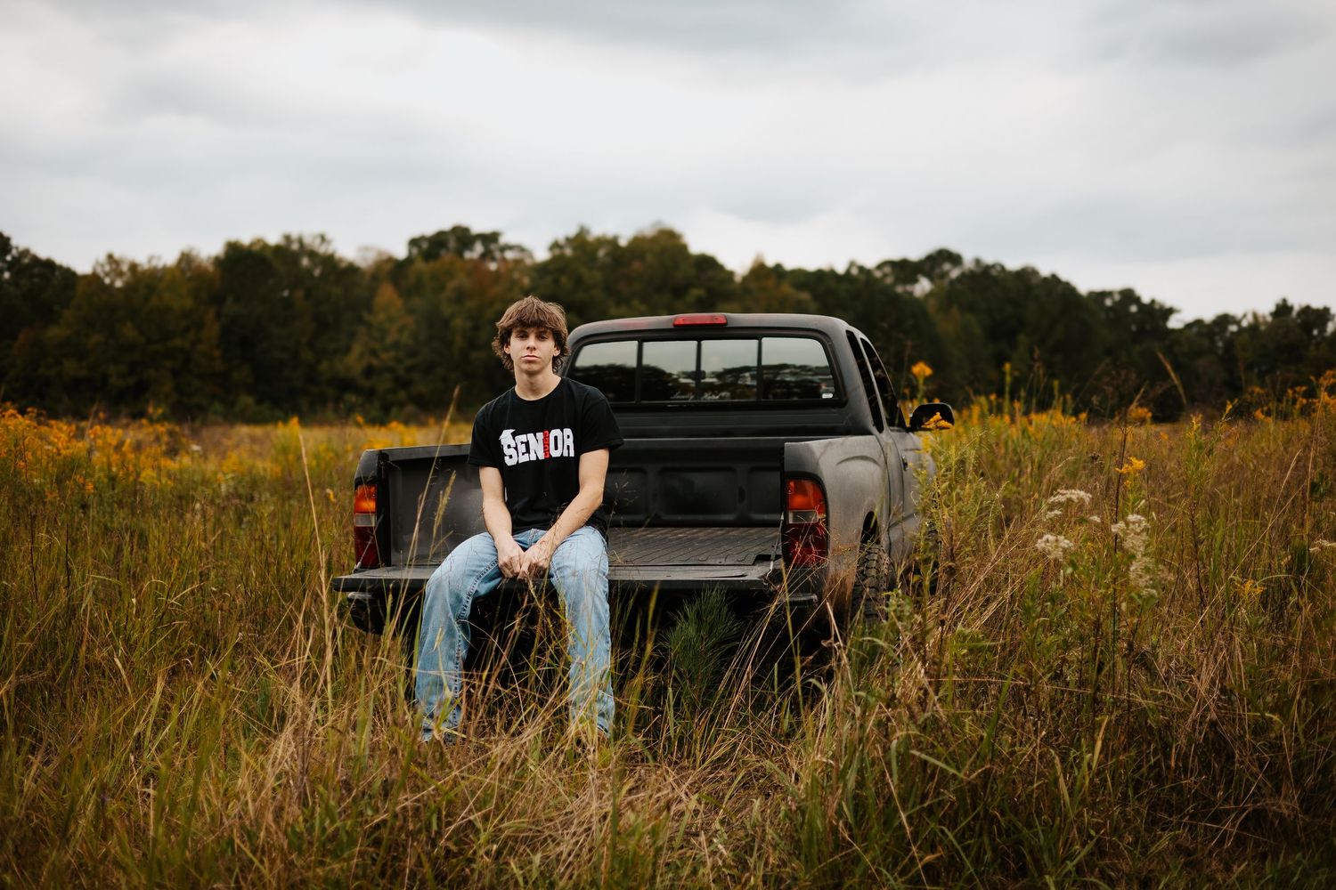 Teen boy in black Senior shirt sitting on tailgate of pickup truck in golden wildflower field.