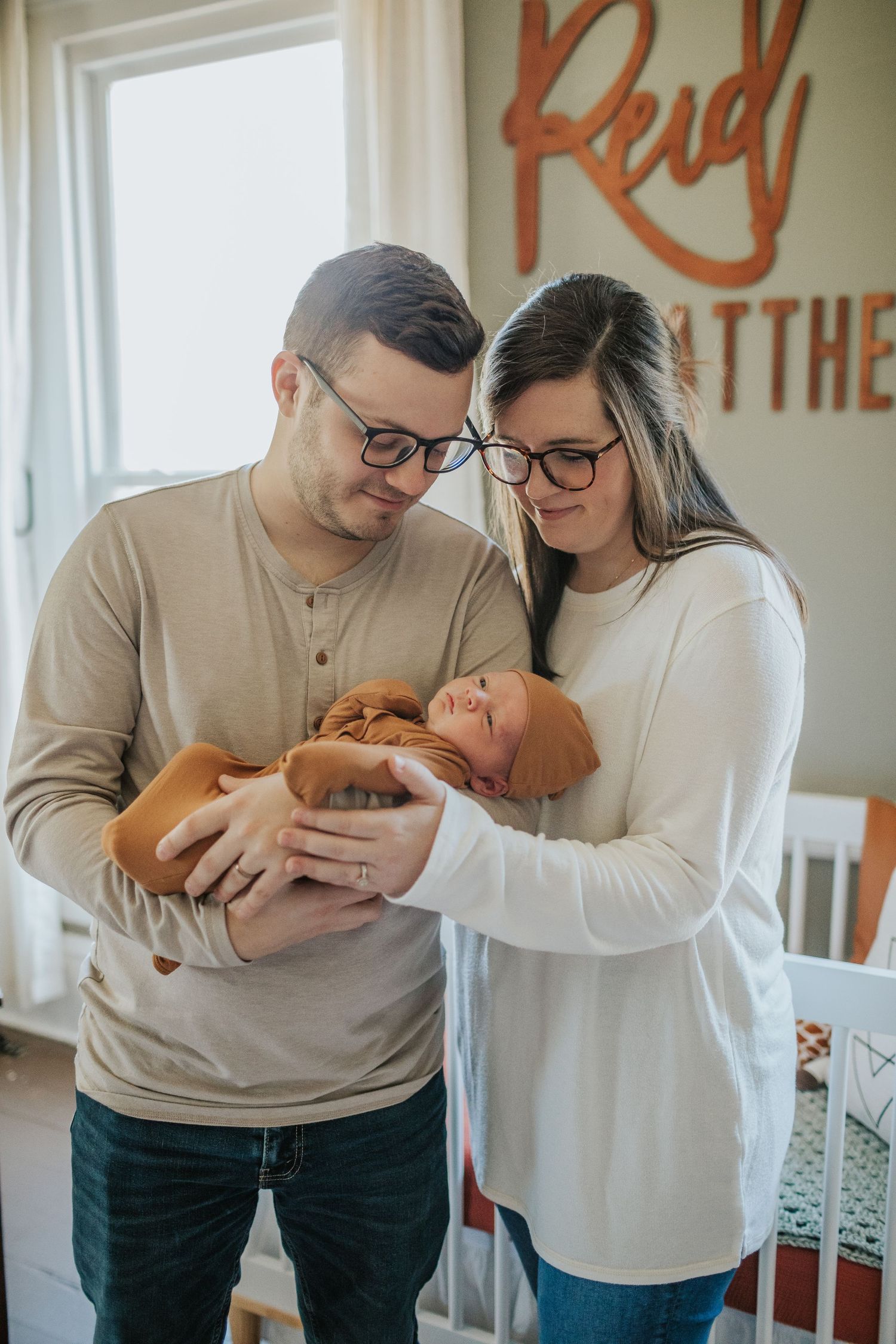 New parents wearing glasses gently hold their sleeping newborn in a decorated nursery room.