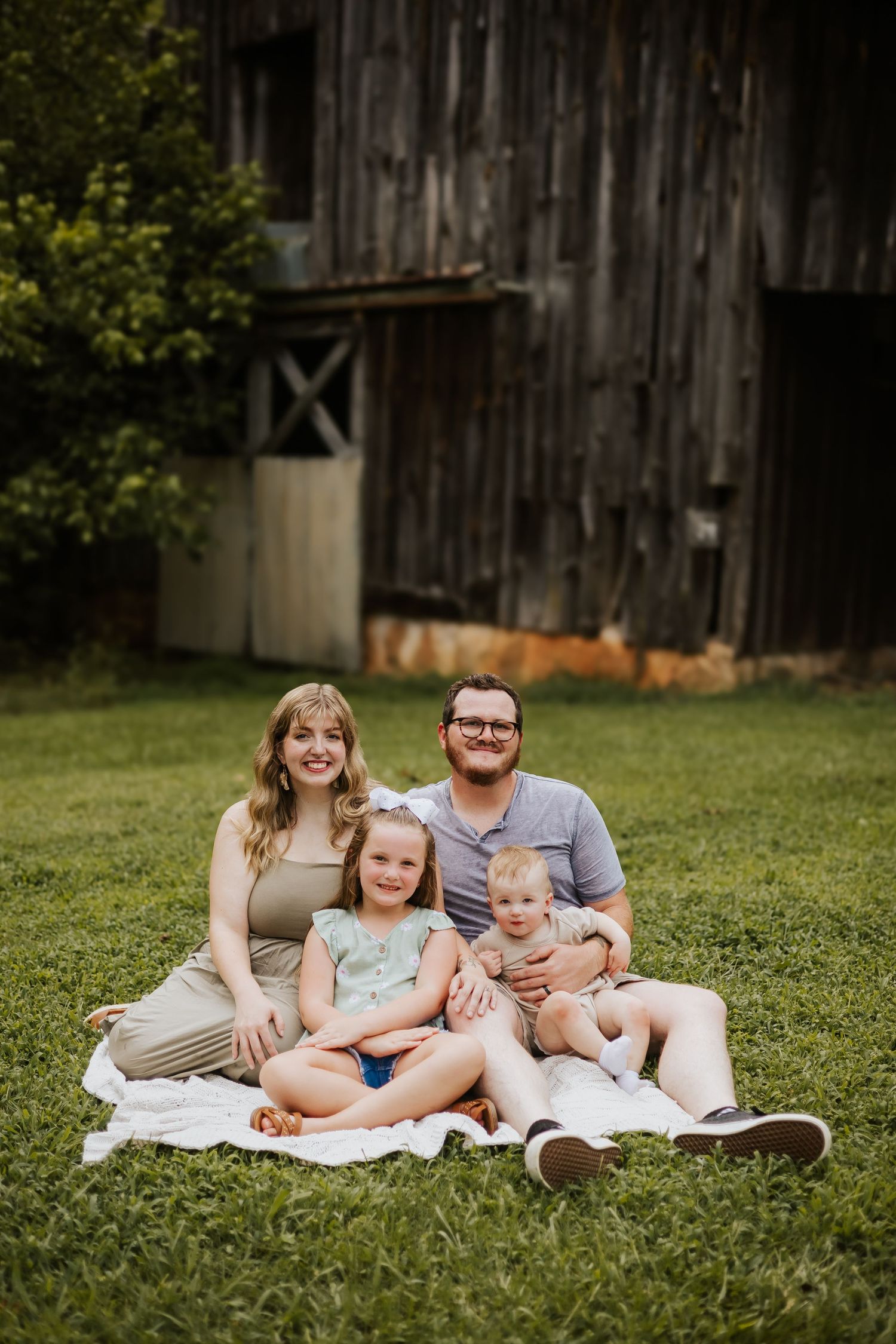 Young family of four sits on a blanket in front of a weathered barn on a sunny day.