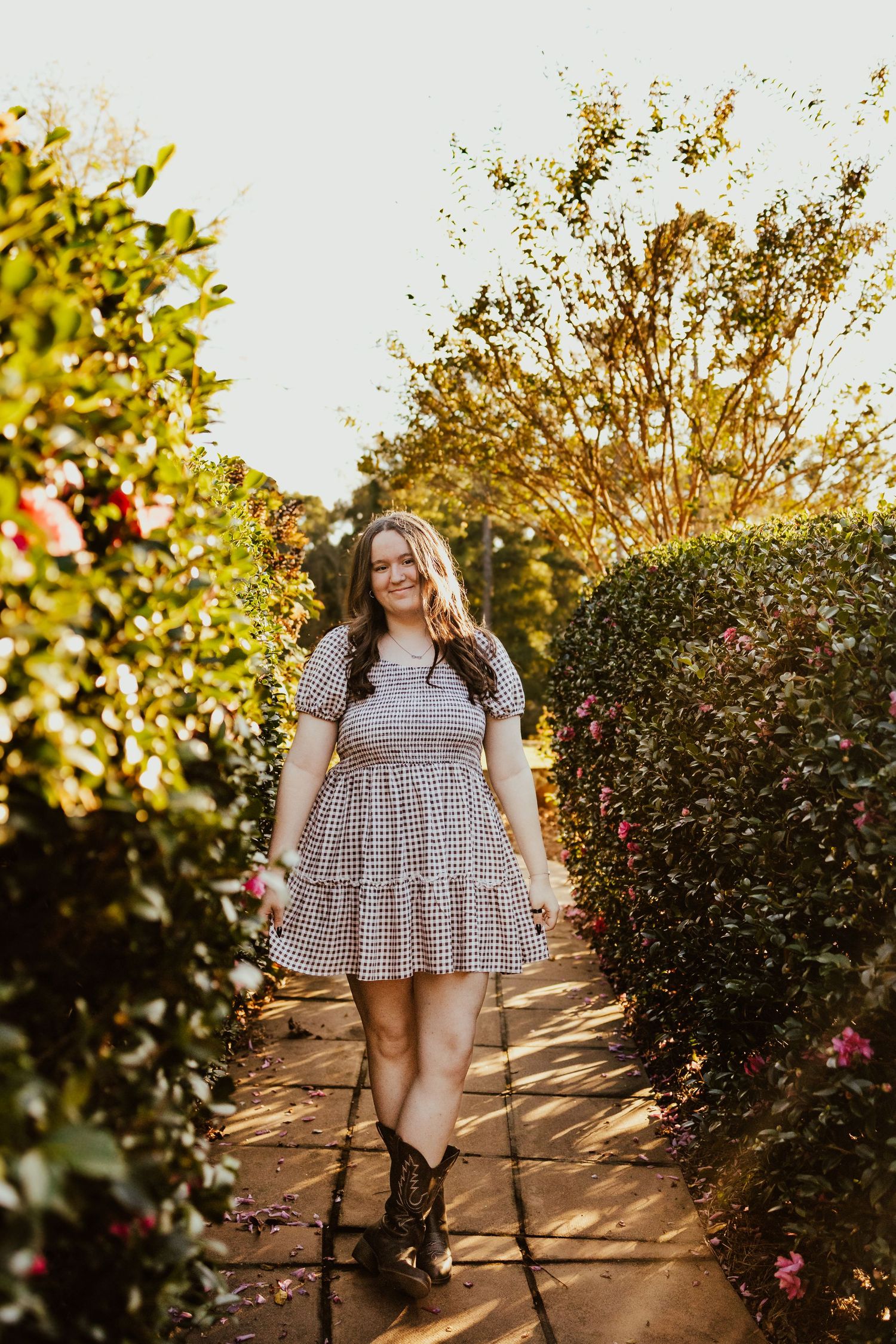 Smiling young woman in gingham dress walking through garden path lined with blooming bushes.