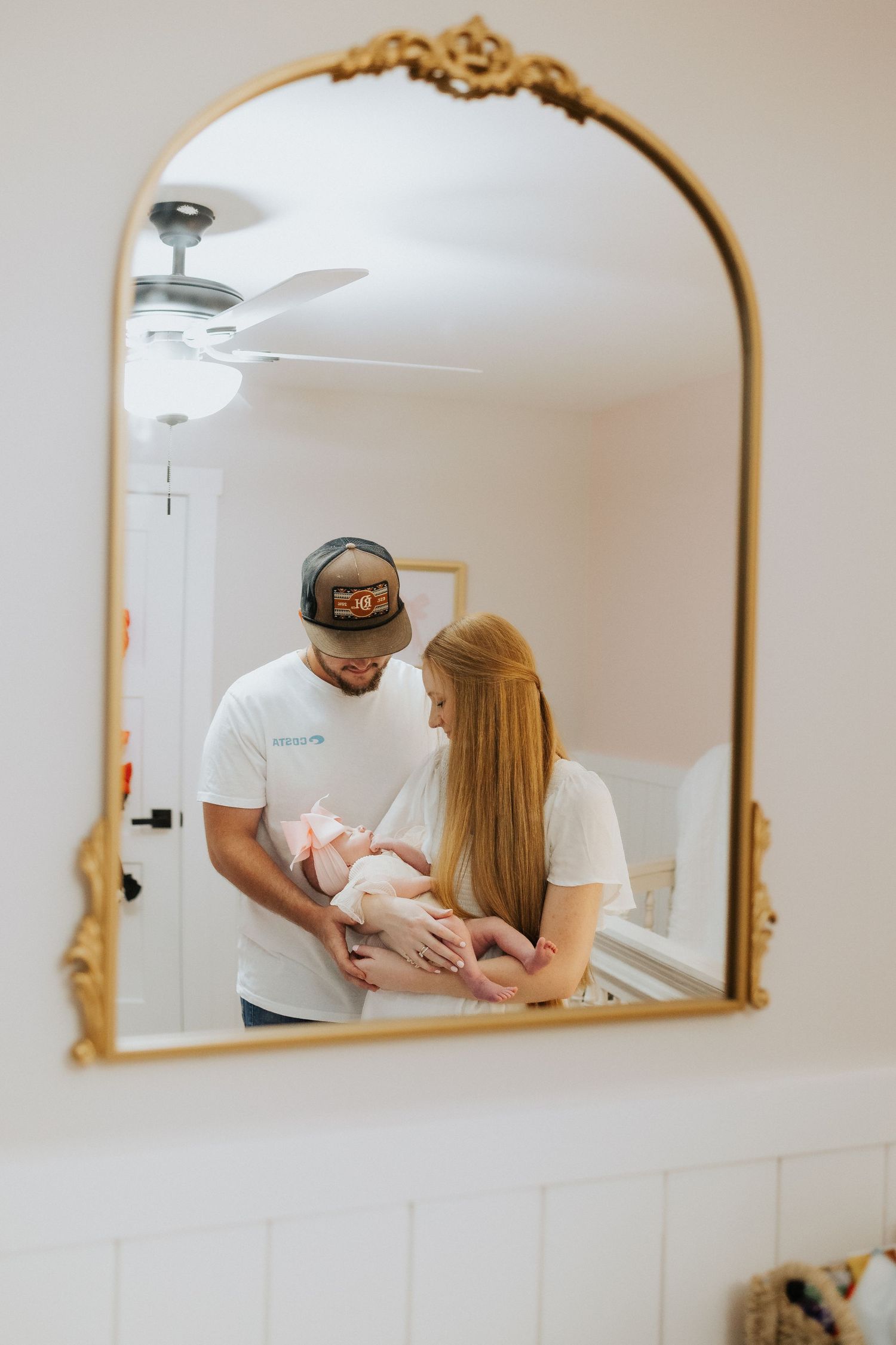 Couple holding newborn baby girl reflected in ornate gold mirror in a bright white nursery room.