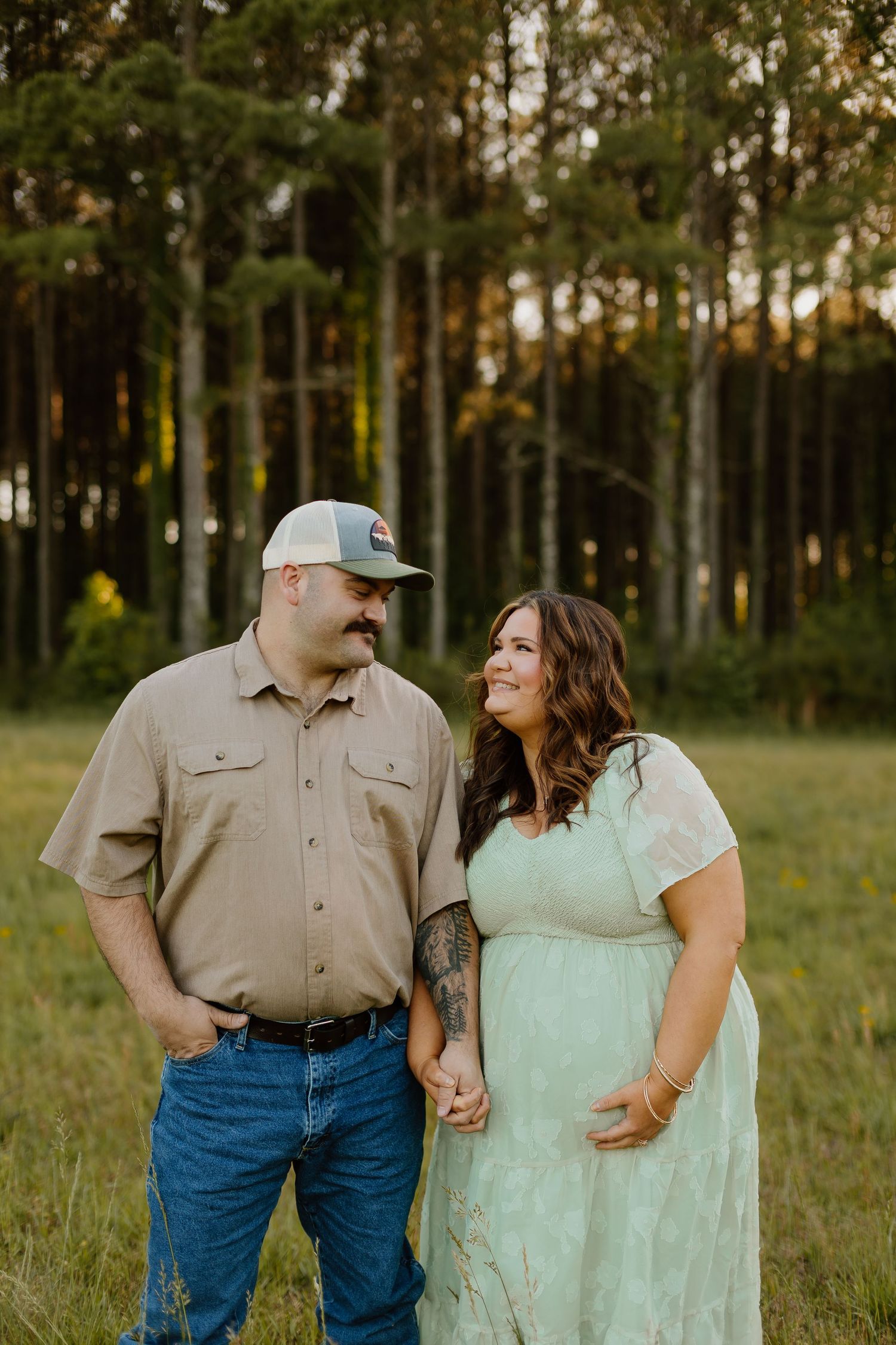 A couple holds hands in a field, the woman in a mint dress cradling her baby bump, with pine trees in the background.