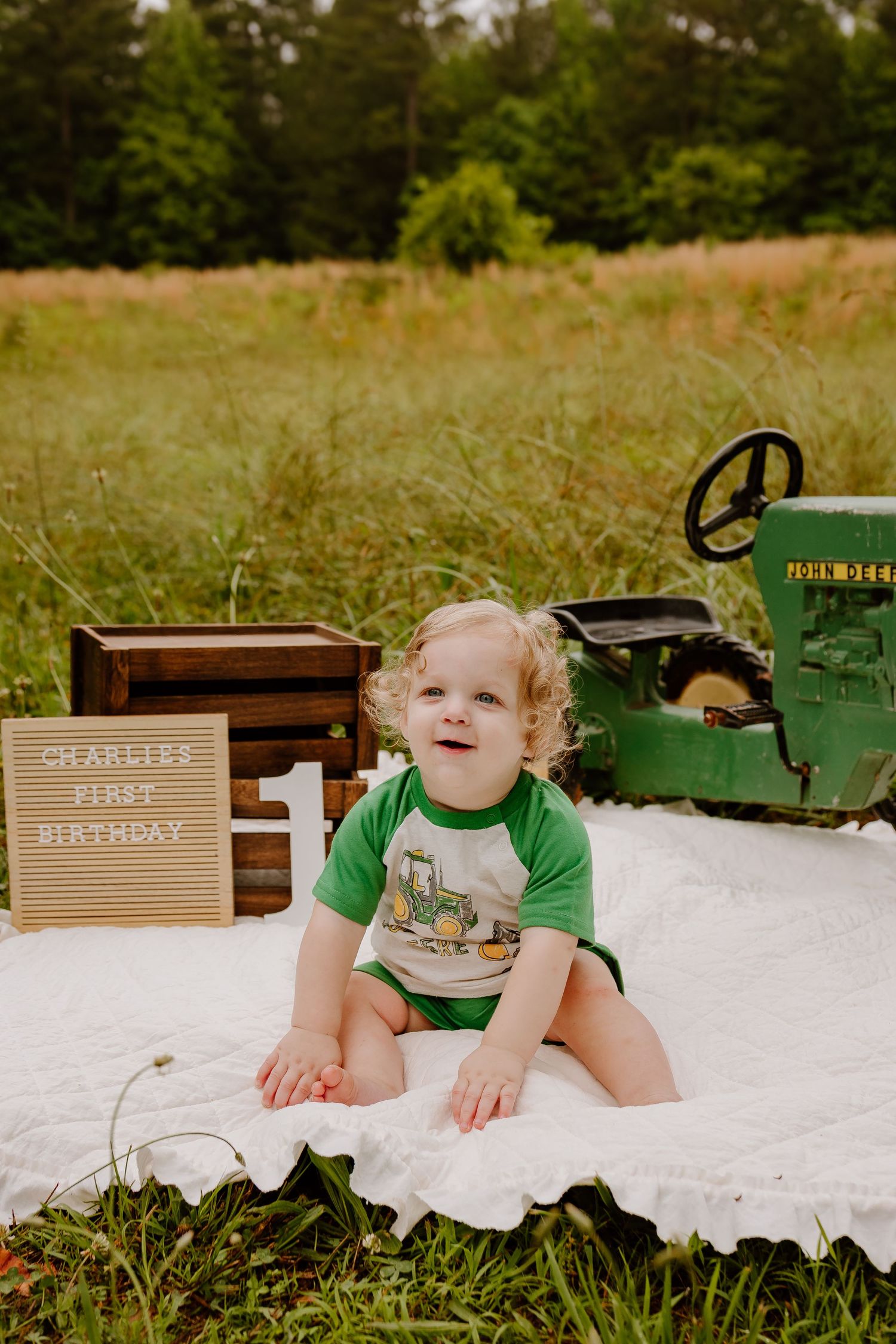 Toddler boy sitting on white blanket in field celebrating Charlie's first birthday with John Deere tractor theme.