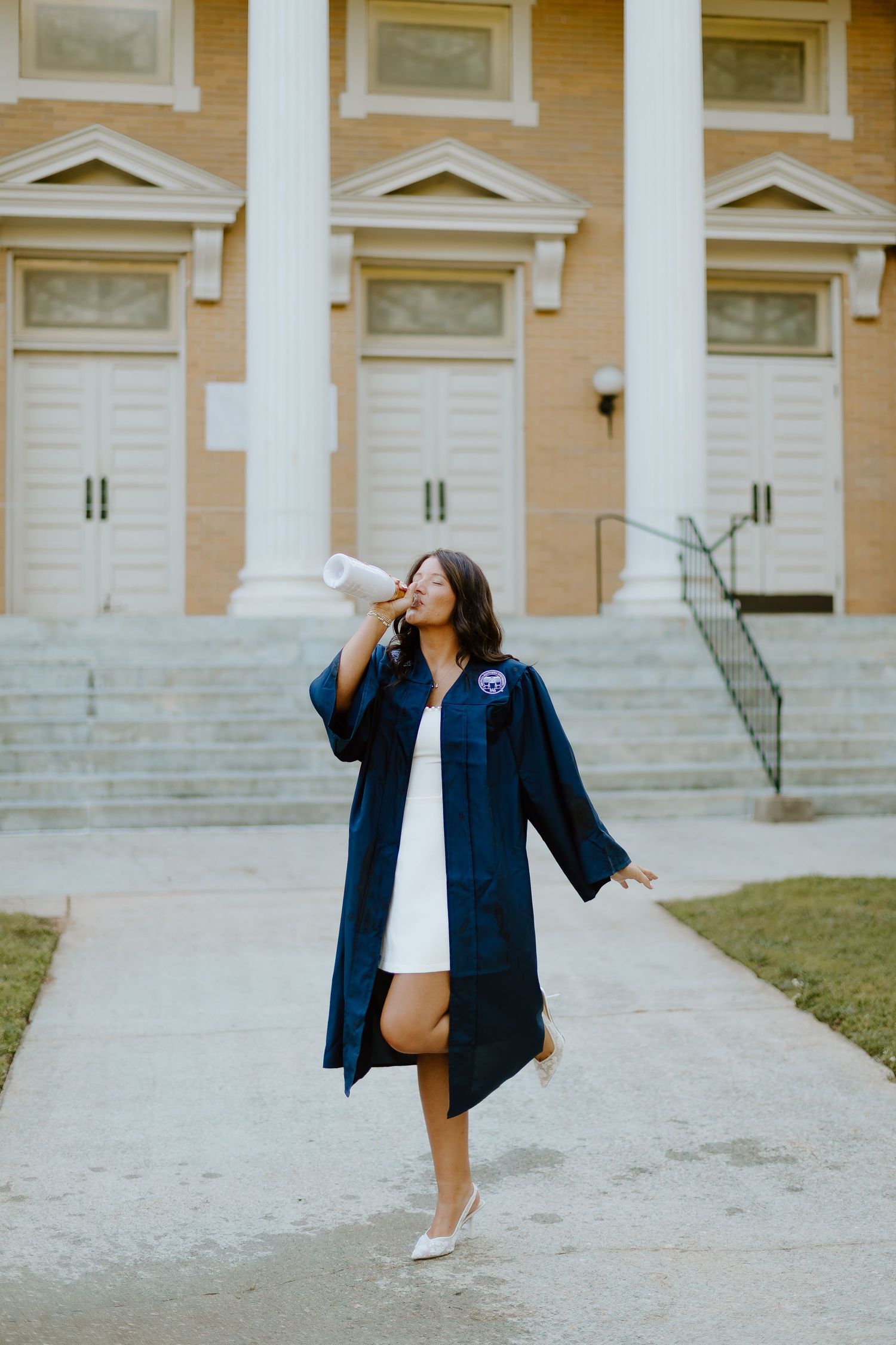 Joyful graduate in navy gown celebrating outside brick university building with white columns.
