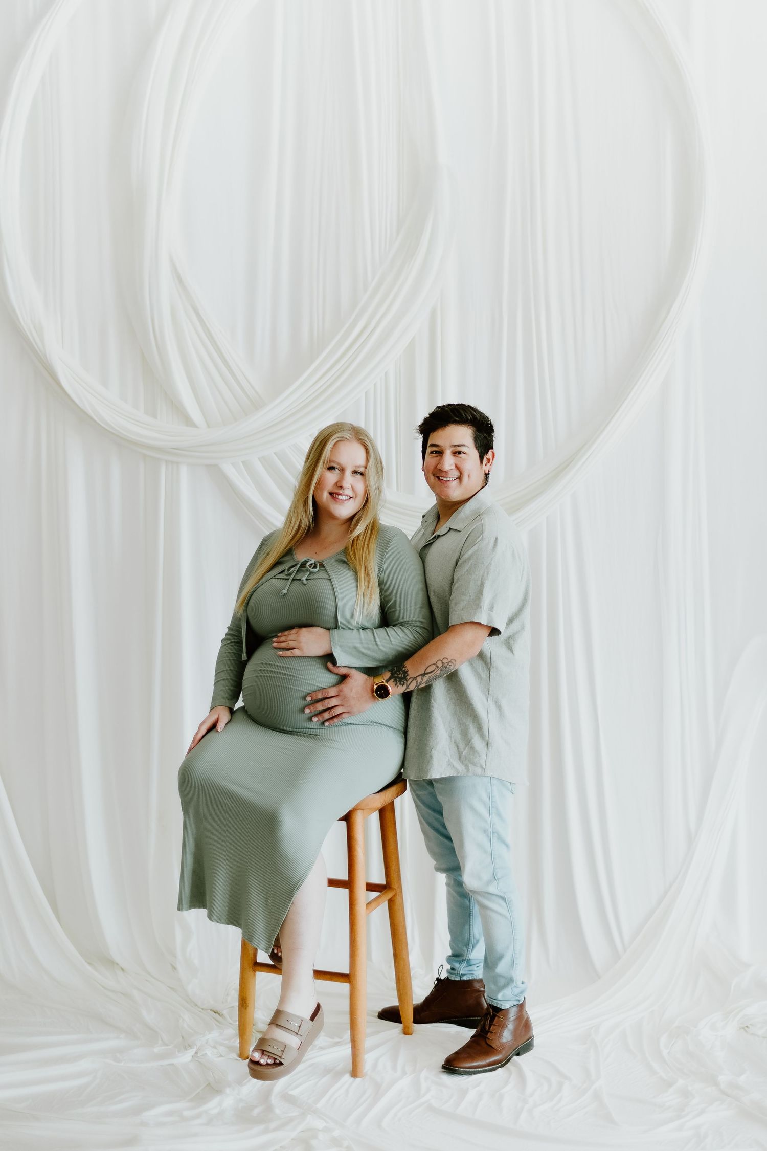 Smiling pregnant woman in sage dress sits on stool with partner in bright white draped studio.