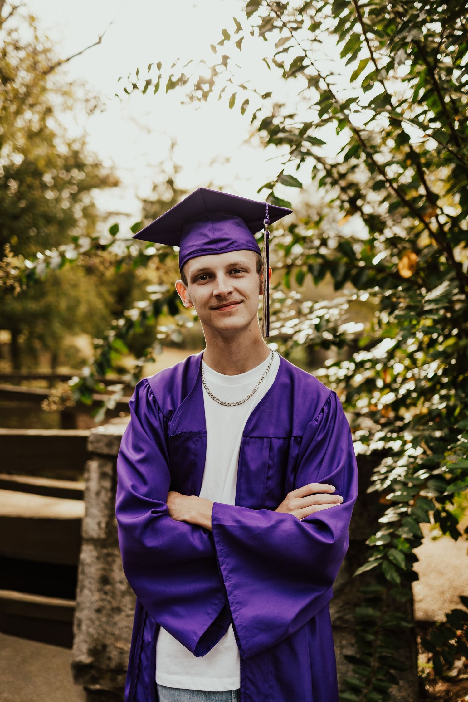 Smiling male graduate in purple cap and gown with arms crossed outdoors among green foliage.