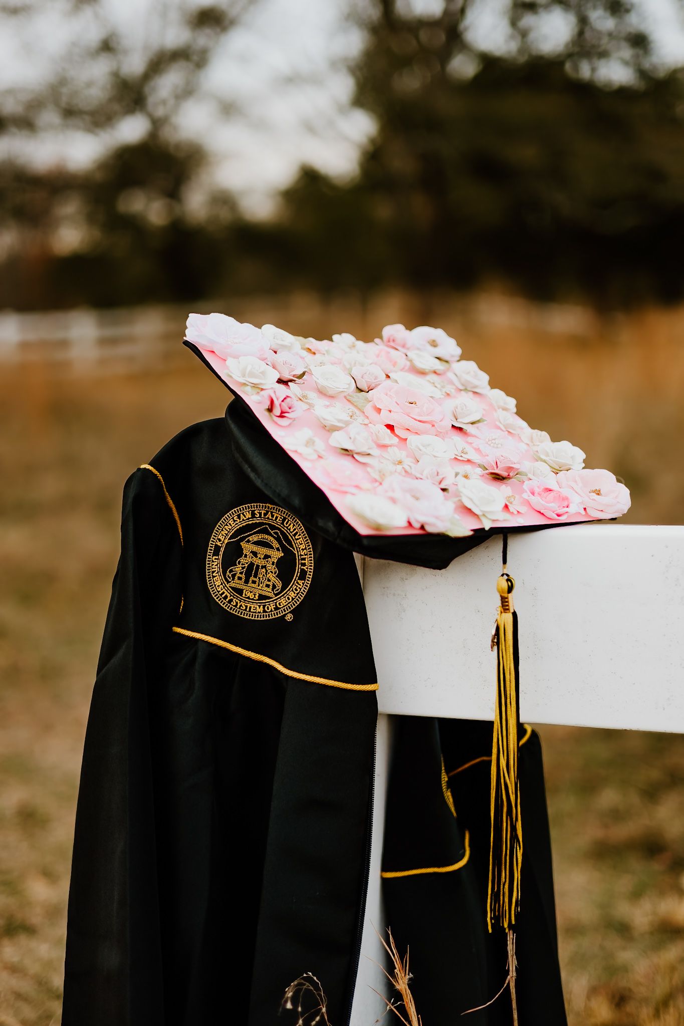 Graduation cap decorated with pink flowers resting on white fence with black gown in field setting.