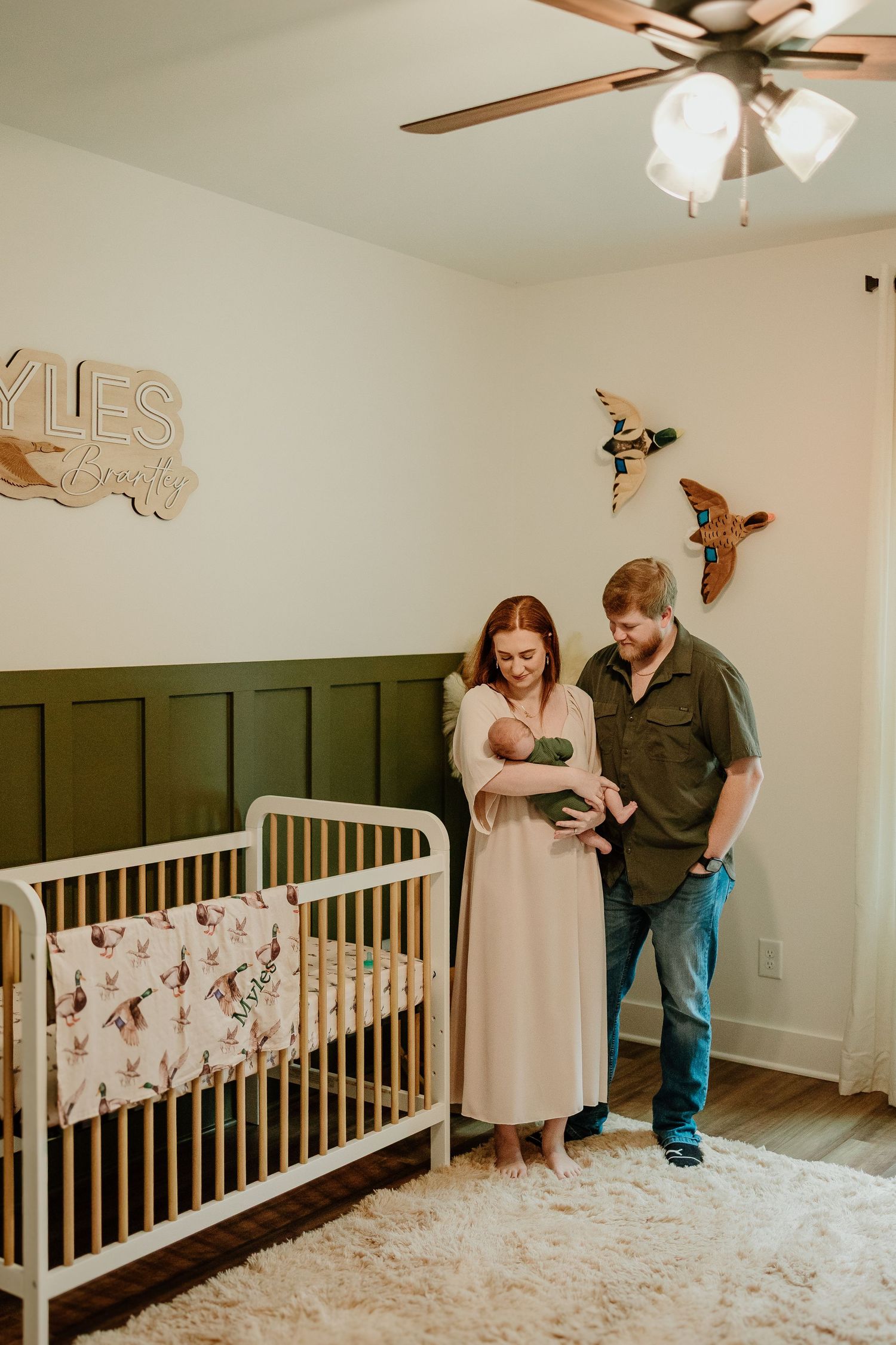 Happy parents holding newborn in bird-themed green nursery with white crib and name sign.