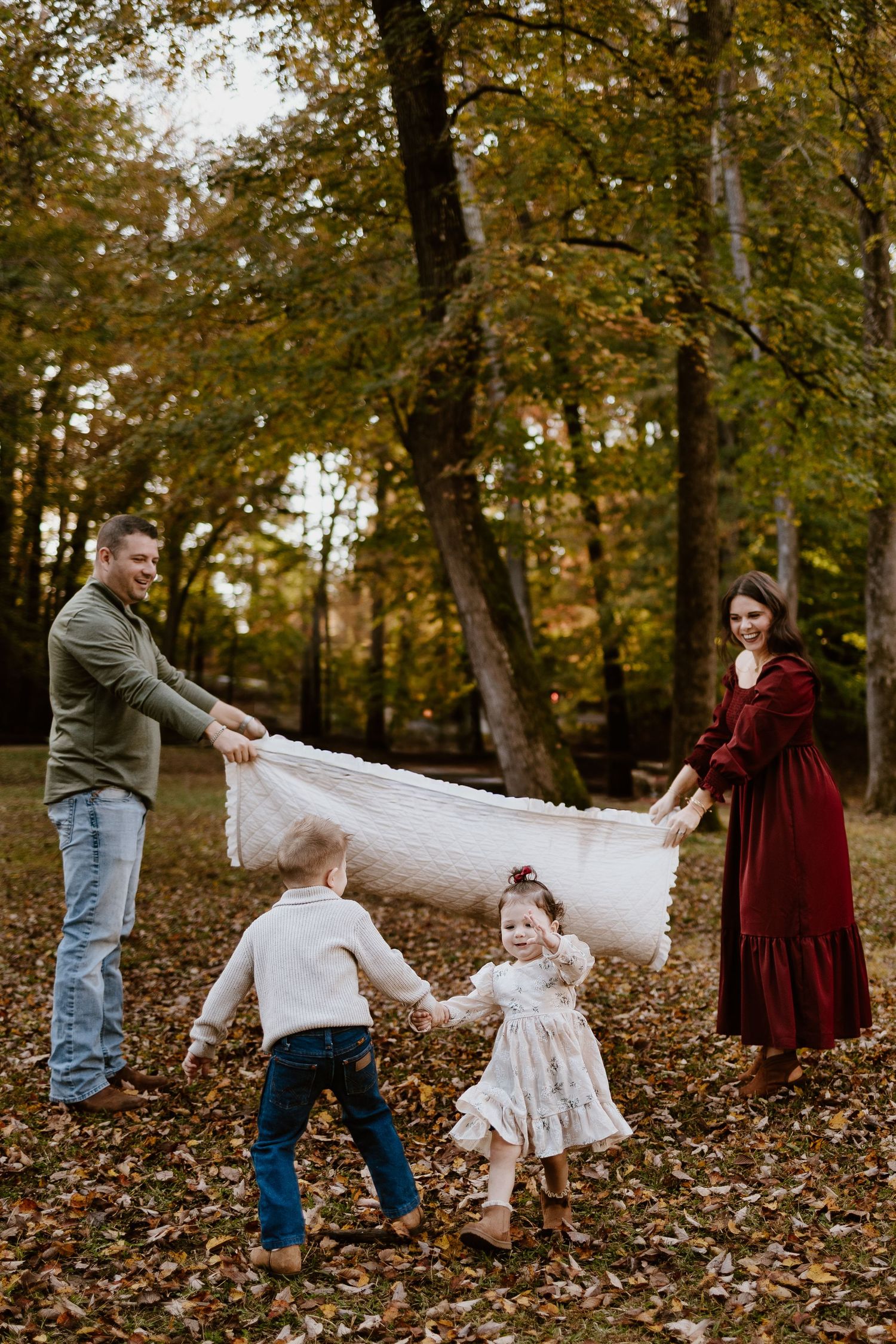 Family of four playing with a white blanket among colorful autumn trees in a forest.