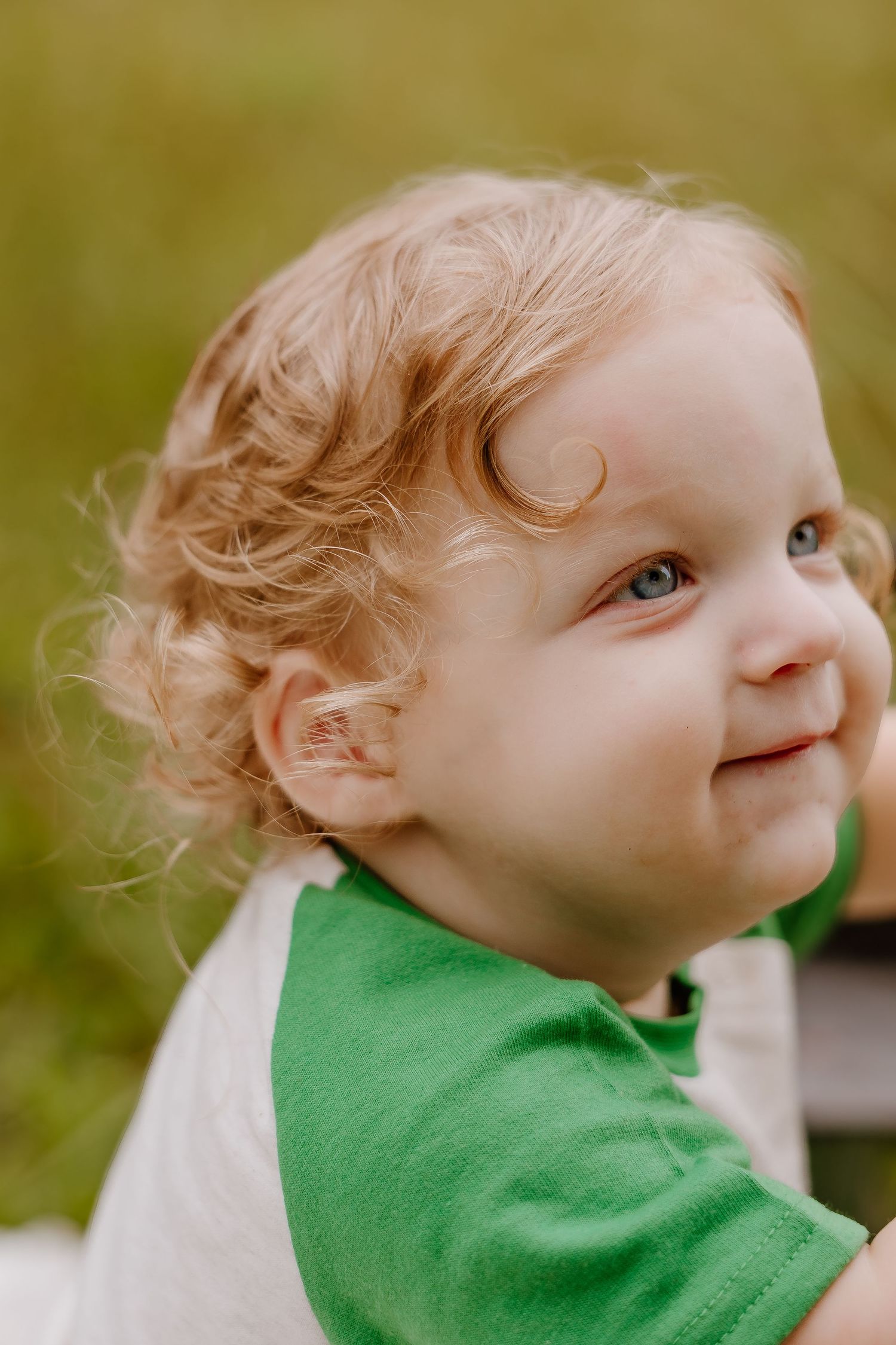 Smiling toddler with curly blonde hair and blue eyes wearing a green and white shirt outdoors.