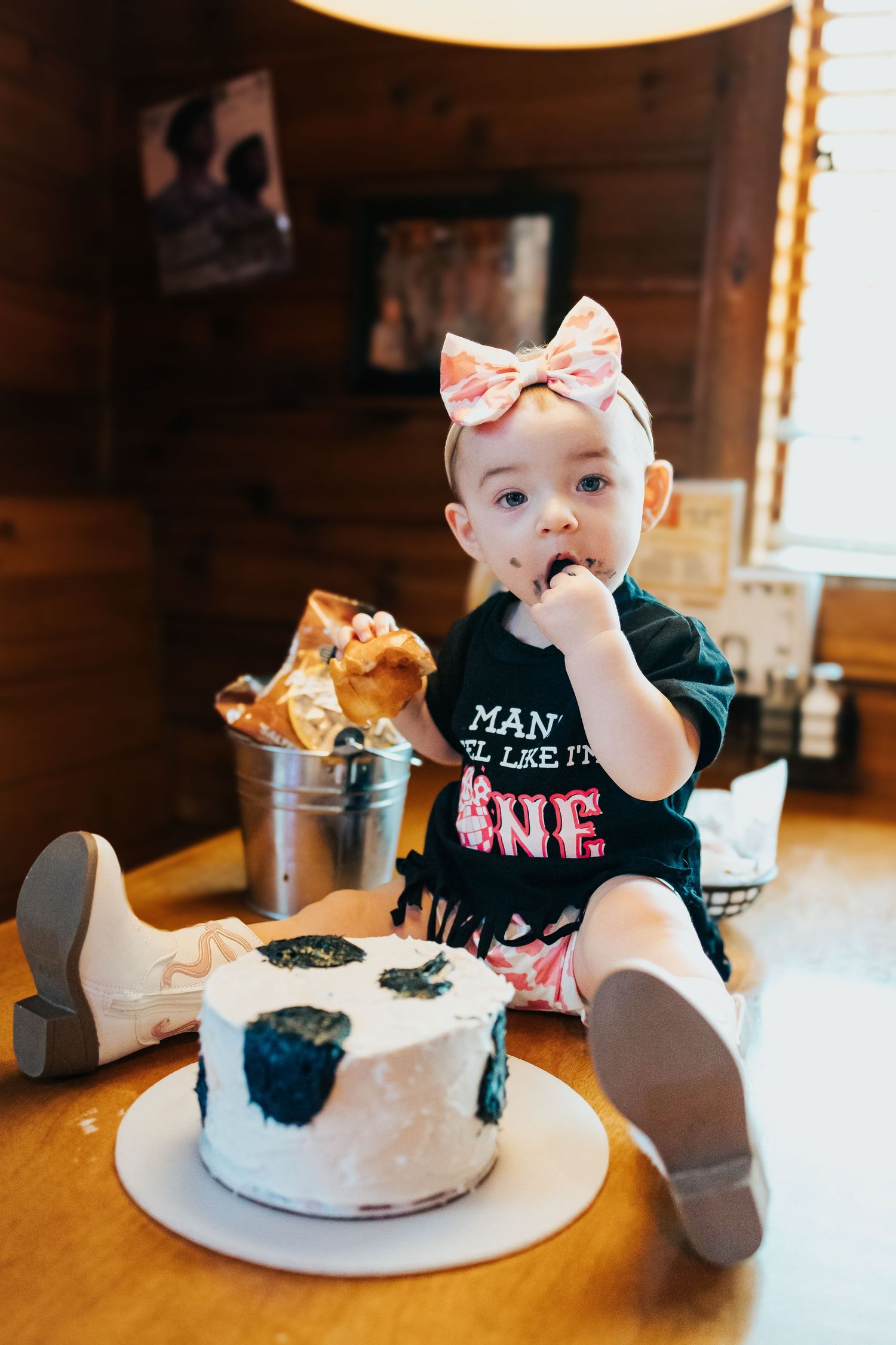 Baby girl in first birthday outfit sits beside smash cake on wooden table during cake smash session.