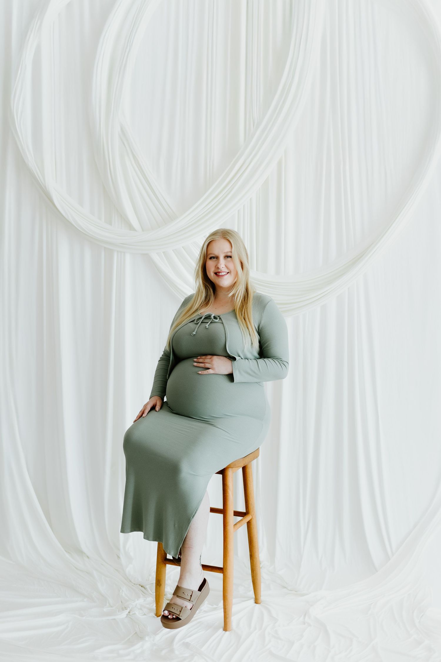Smiling pregnant woman in sage green maternity dress sits on wooden stool in white draped studio.