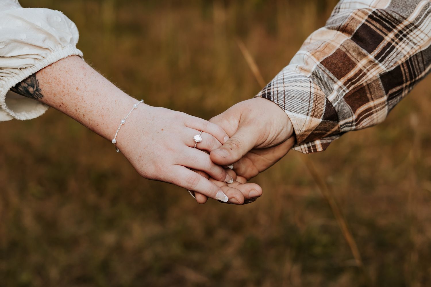 Close-up of couple holding hands showing engagement ring and bracelet in autumn field setting.