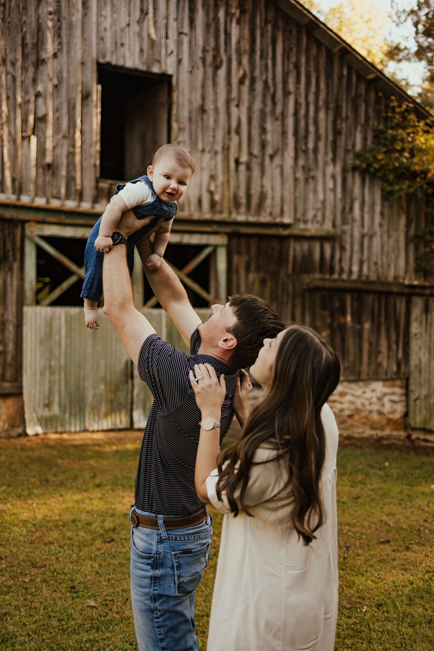 Joyful father lifts laughing baby in the air while mother watches near a rustic barn at sunset.