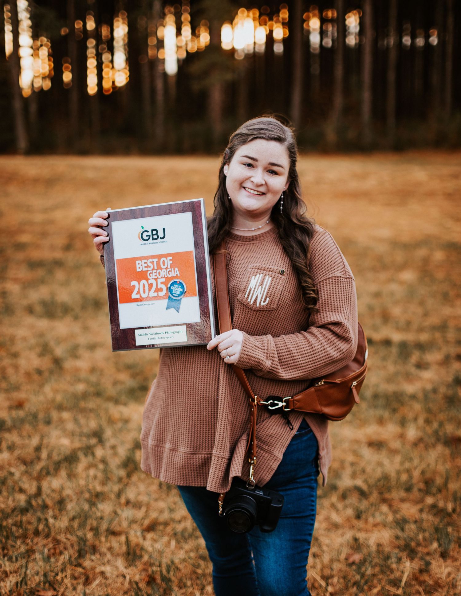 Smiling young woman holding a Best of Georgia 2025 GBJ award in an open field at sunset.