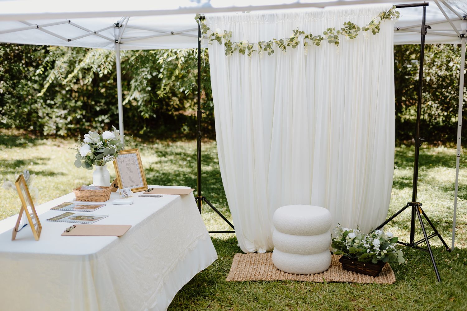 Outdoor wedding setup with white drape backdrop, floral garland, draped table, and stacked ottoman on woven rug.