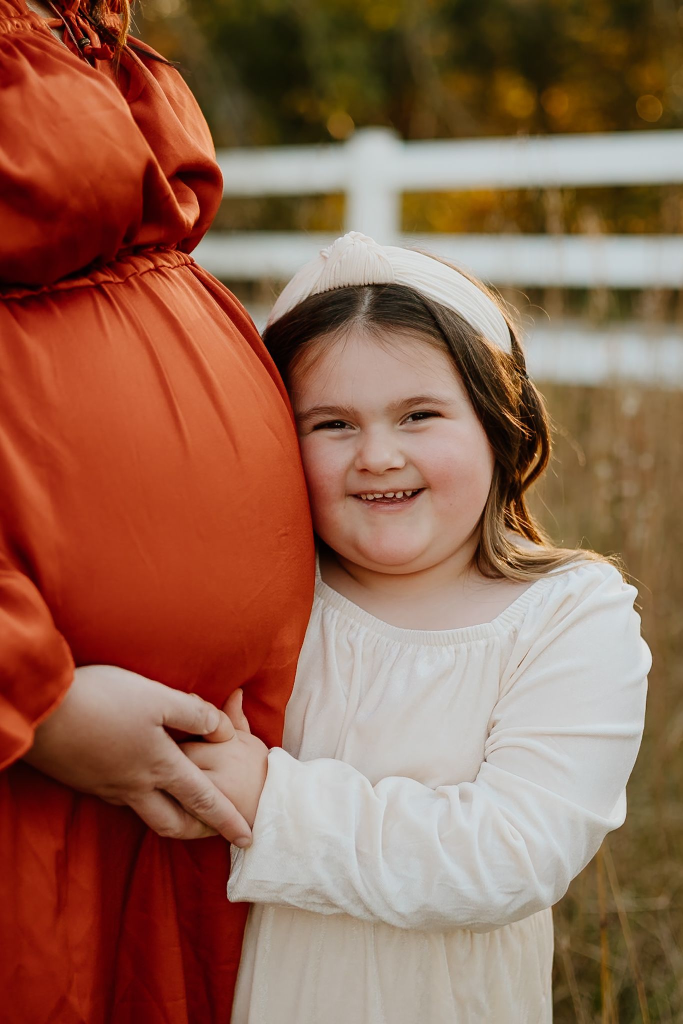 A young girl in a white dress hugs her mother's pregnant belly outdoors during a fall maternity photo session.
