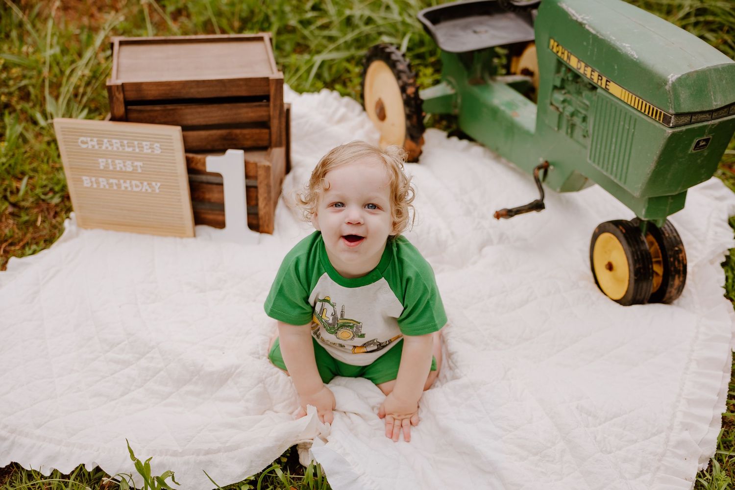 Smiling toddler in green shirt sits on white blanket beside John Deere tractor toy and first birthday sign outdoors.