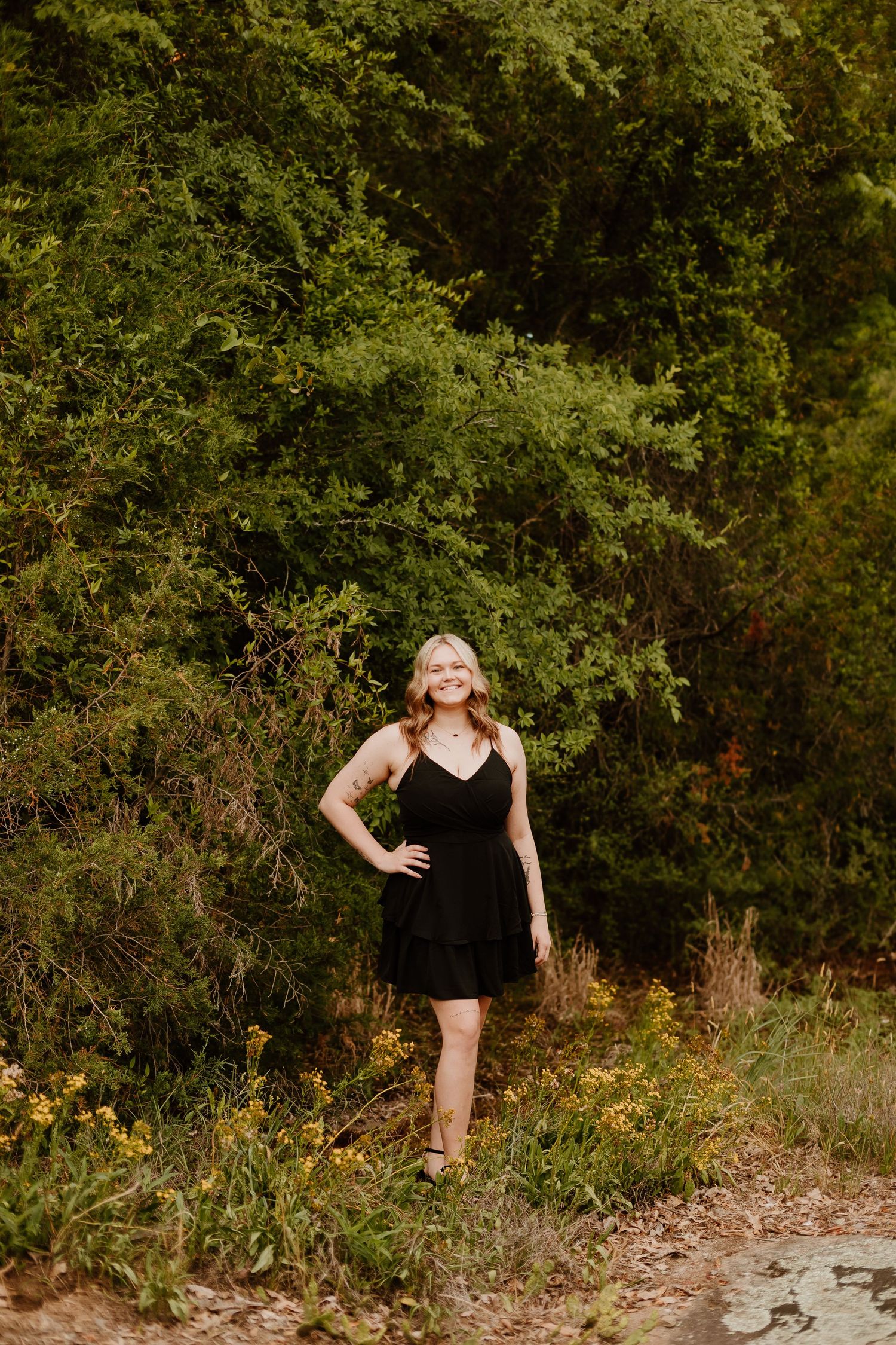 Woman in black dress smiling outdoors among wildflowers and lush green trees on a nature trail.