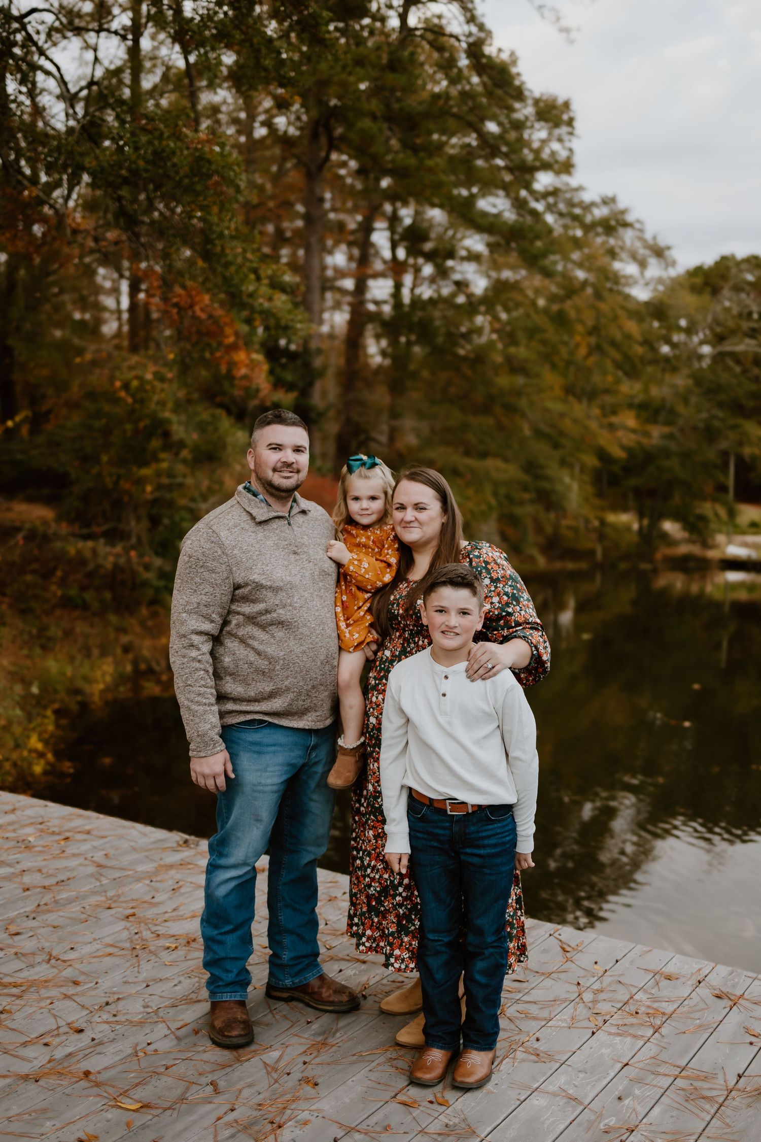 Family of four posing on a wooden dock beside a lake surrounded by colorful fall foliage.