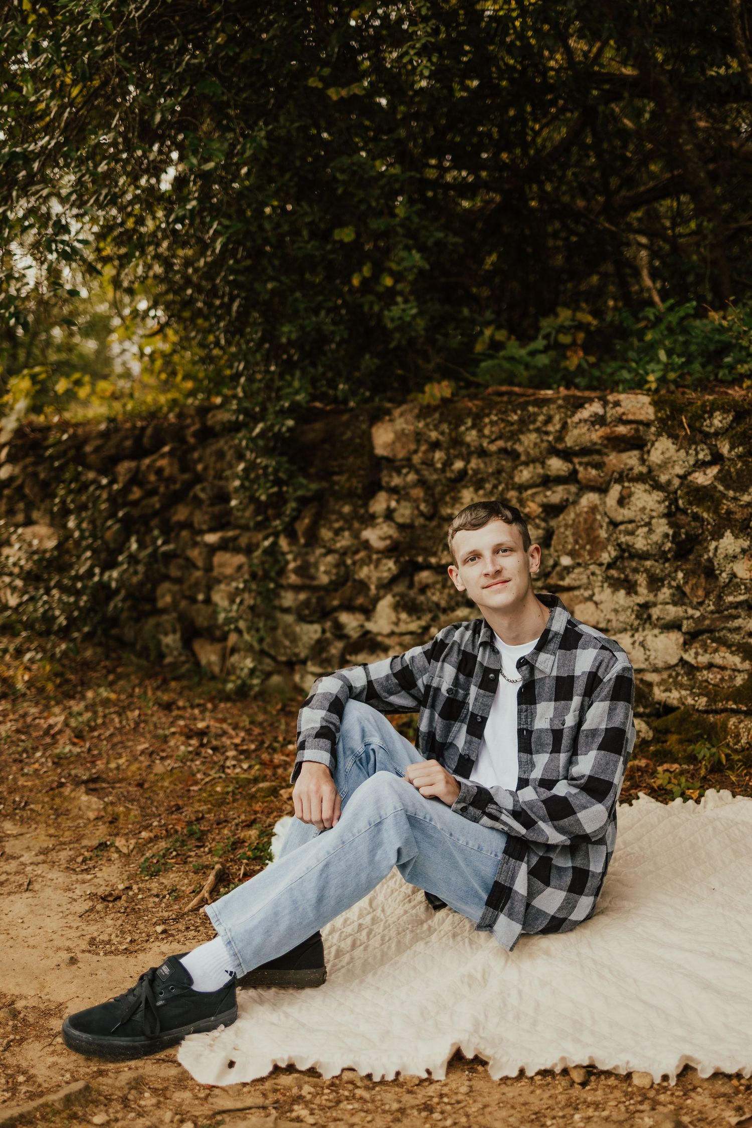 Young man in plaid shirt and jeans sitting on white rug against rustic stone wall outdoors.