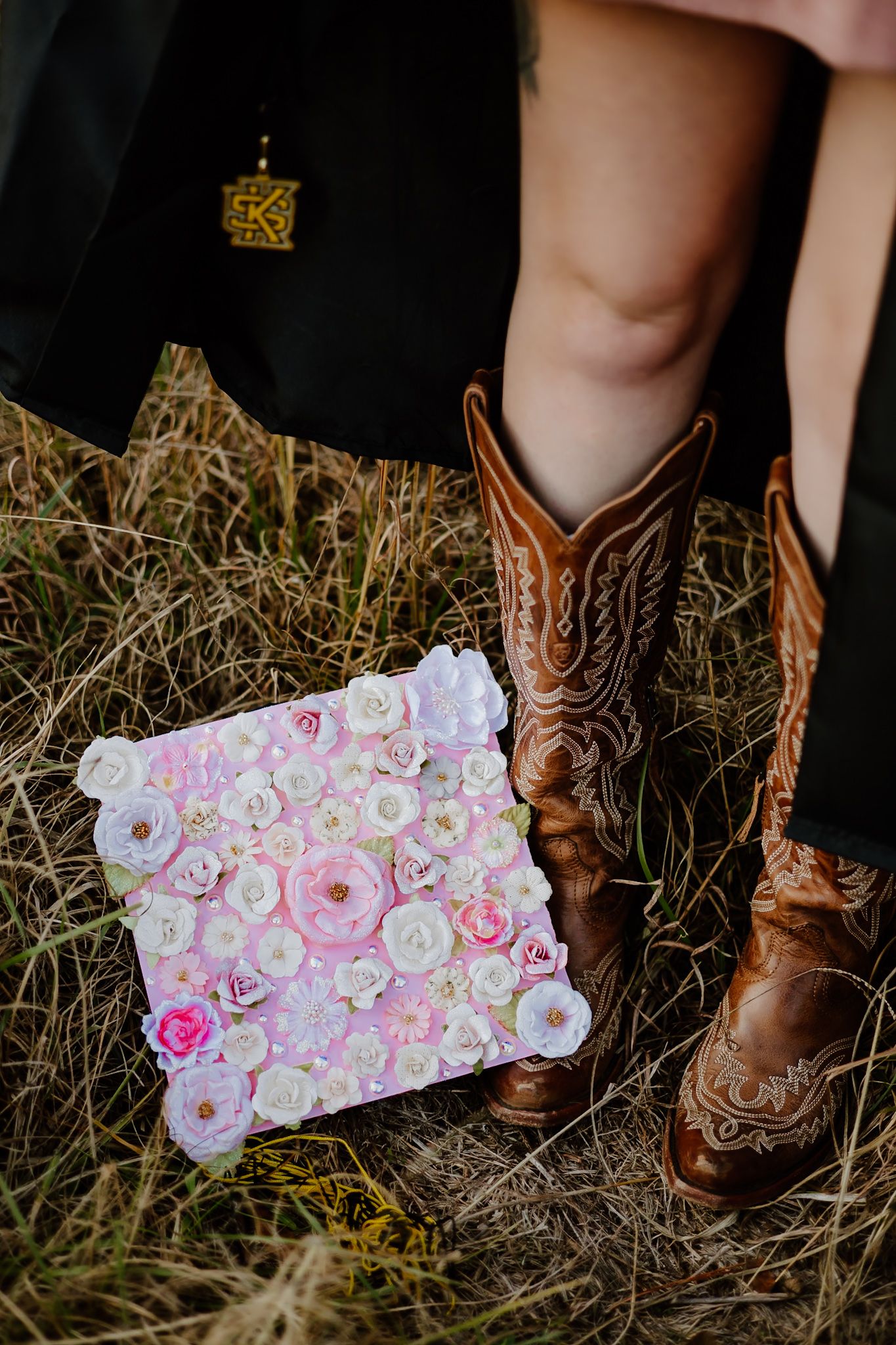 Close-up of floral decorated graduation cap beside cowboy boots in dry grass field.