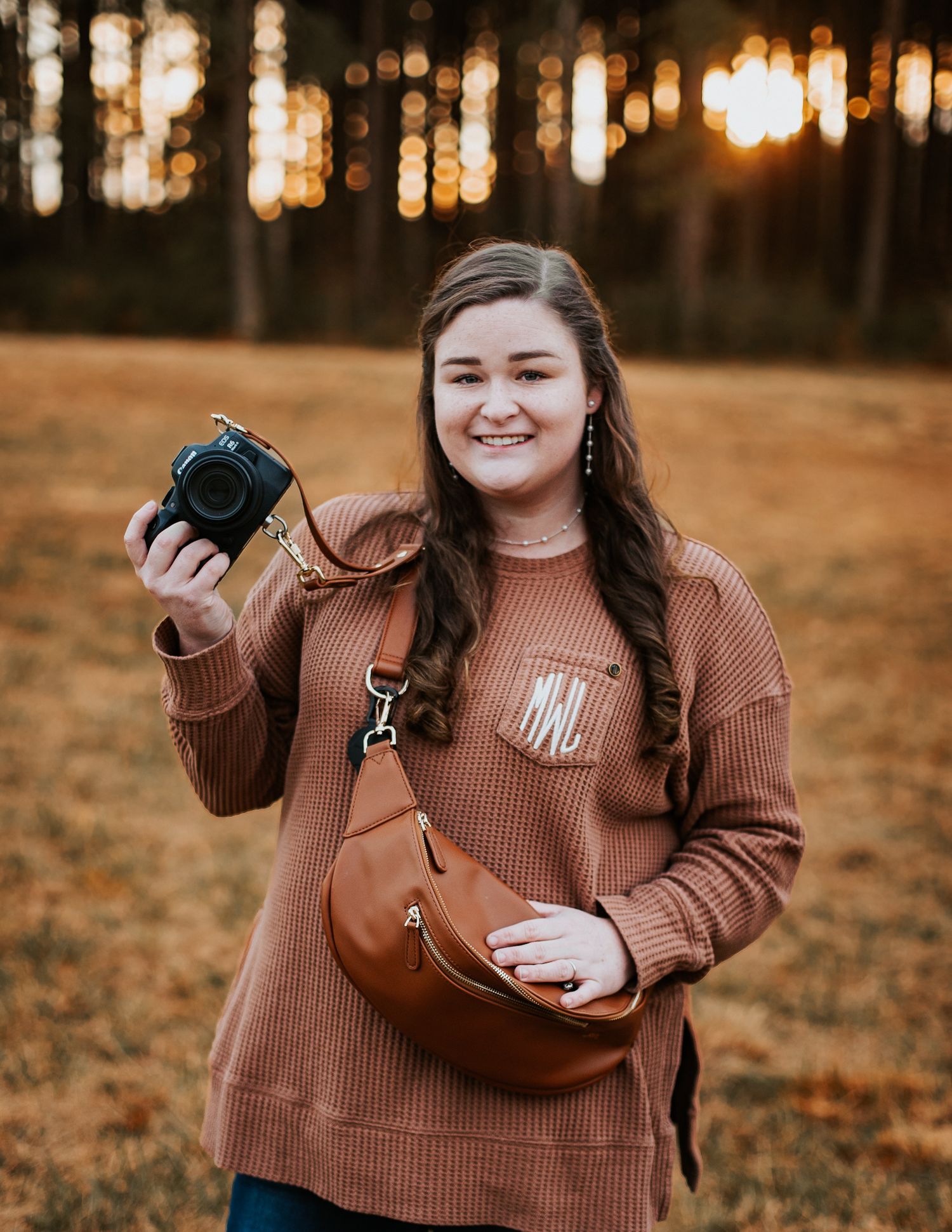 Young female photographer smiling in a russet sweater holding a camera outdoors at golden hour.