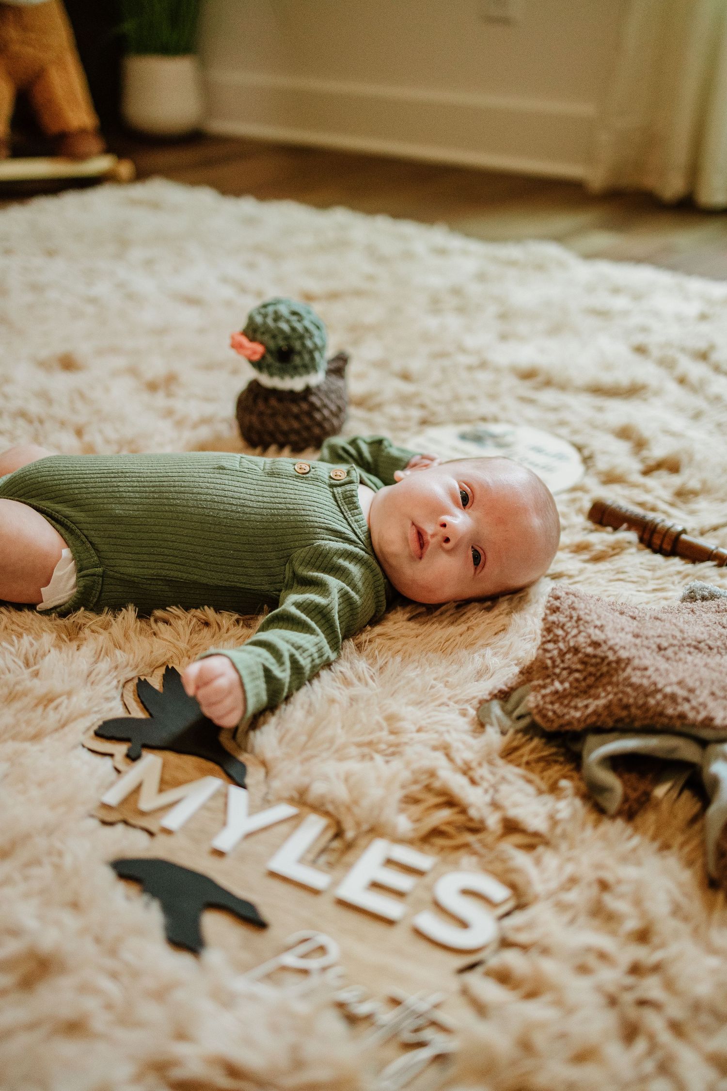 Newborn baby Myles in green onesie lying on fluffy rug surrounded by name letters and toys.