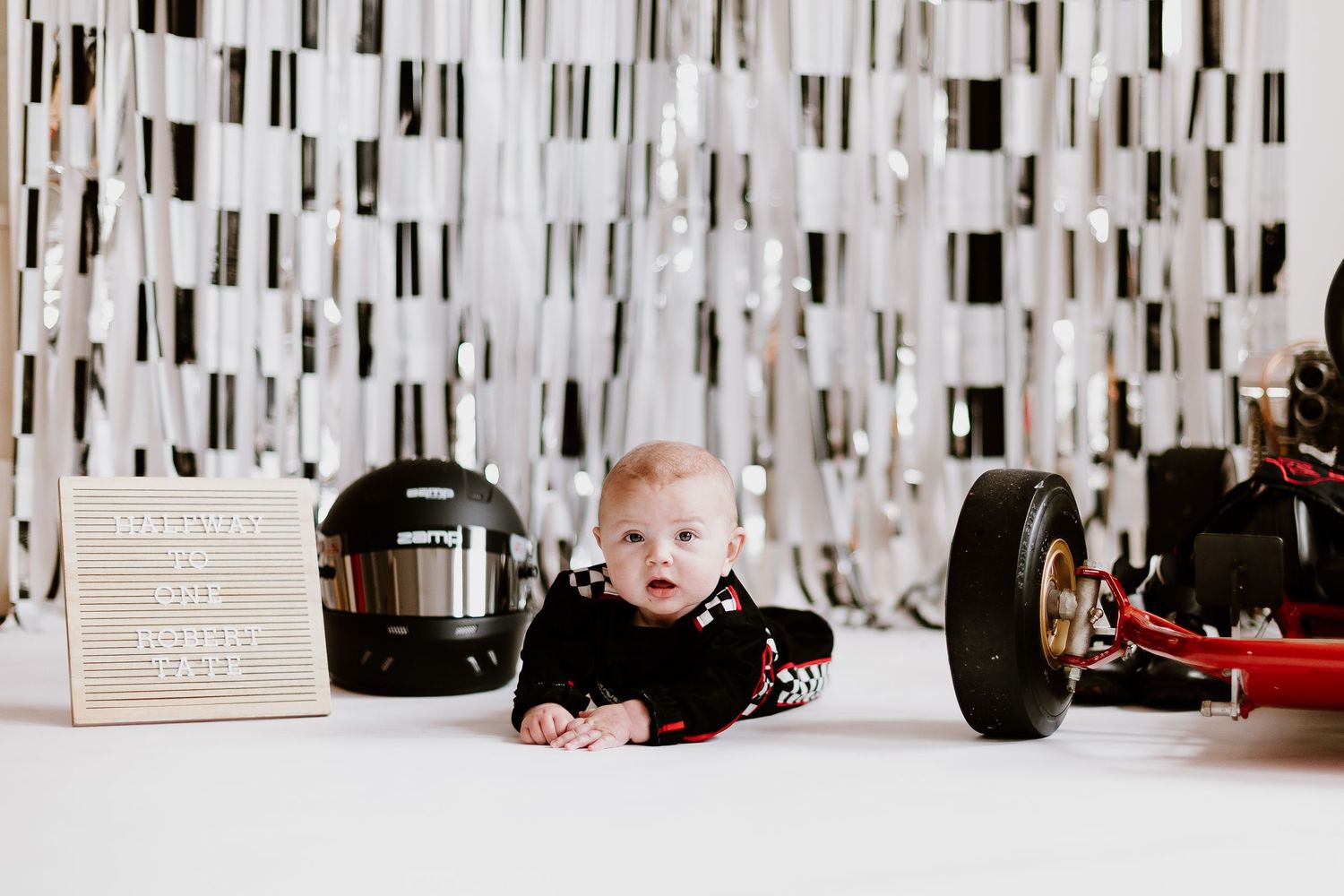 Baby in racing outfit lies on floor beside go-kart and helmet for half-birthday race themed photoshoot.