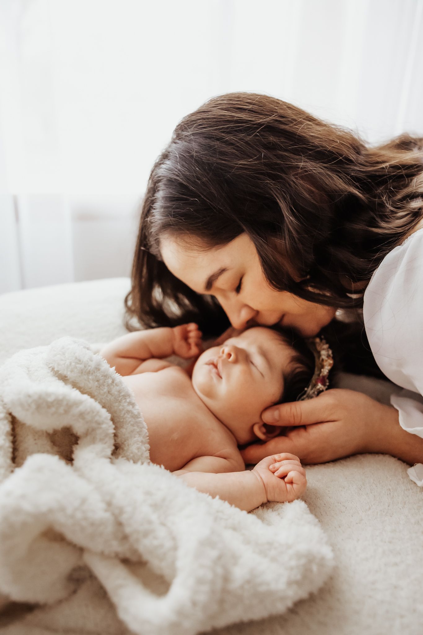A tender moment as a dark-haired mother gently kisses her sleeping newborn baby lying on a soft white blanket.