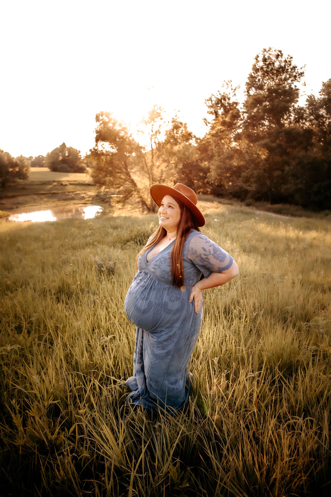 A pregnant woman in a blue lace gown and brown hat glows in golden sunlight in a green field.