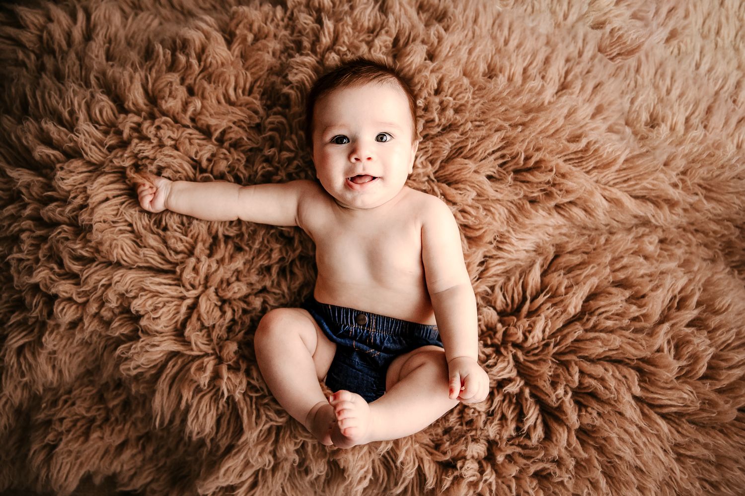 Happy baby lying on fluffy brown rug, looking up with bright curious eyes.
