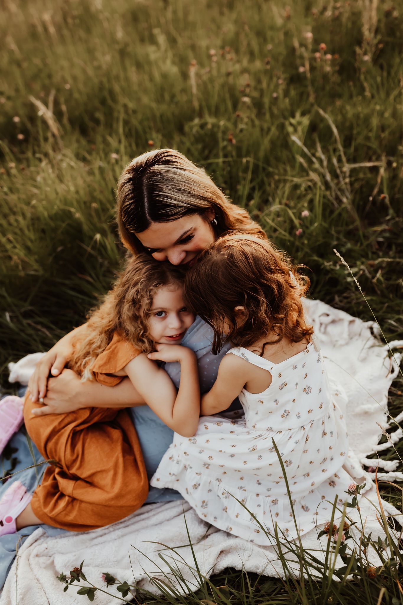 A loving mother sits in a green meadow embracing her two young daughters in a warm golden light setting.